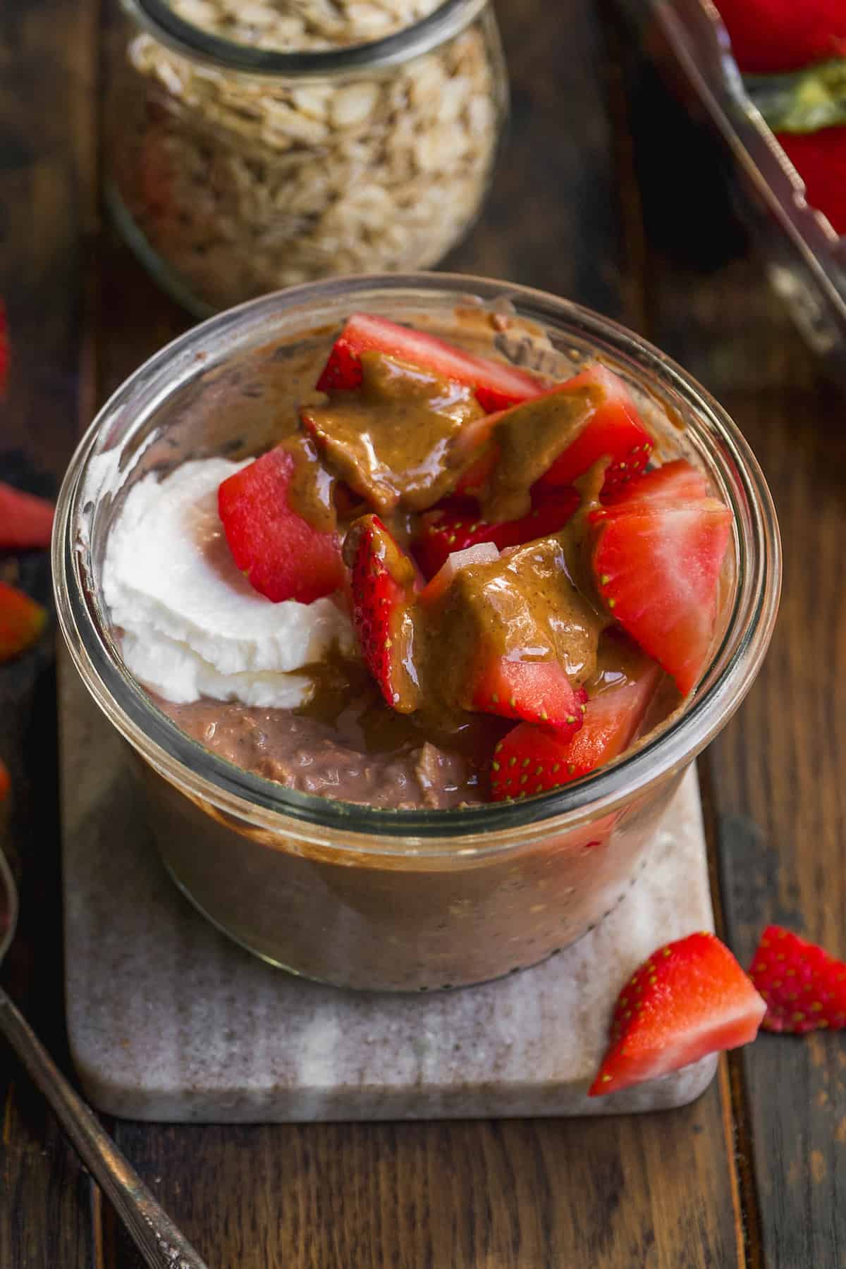 A glass jar filled with chocolate overnight oats, topped with sliced strawberries, a dollop of yogurt, and a drizzle of nut butter, sits on a small marble board with a few strawberry pieces beside it.