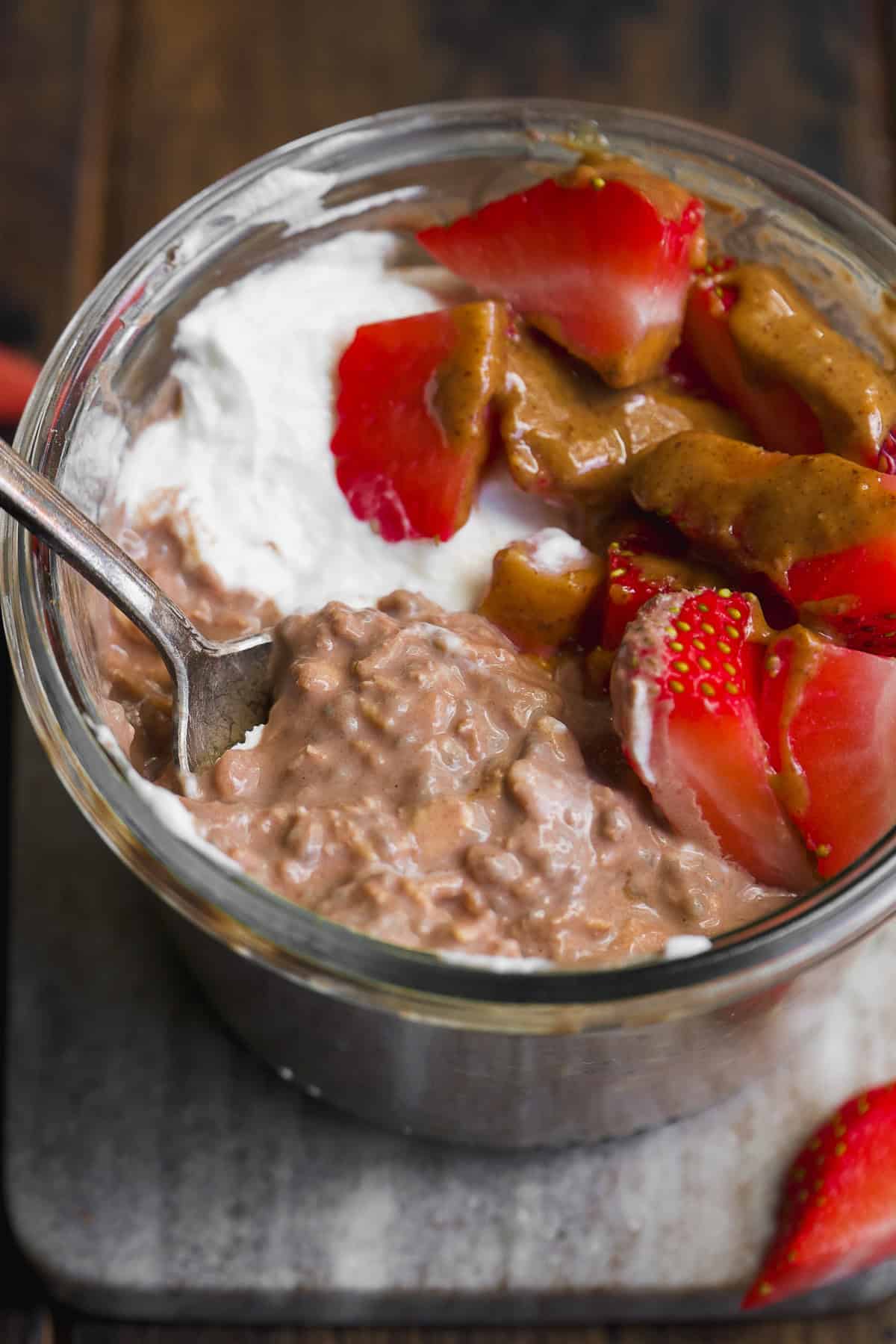 A glass jar filled with chocolate overnight oats, topped with sliced strawberries, a dollop of whipped cream, and a drizzle of nut butter, with a spoon inside the jar.