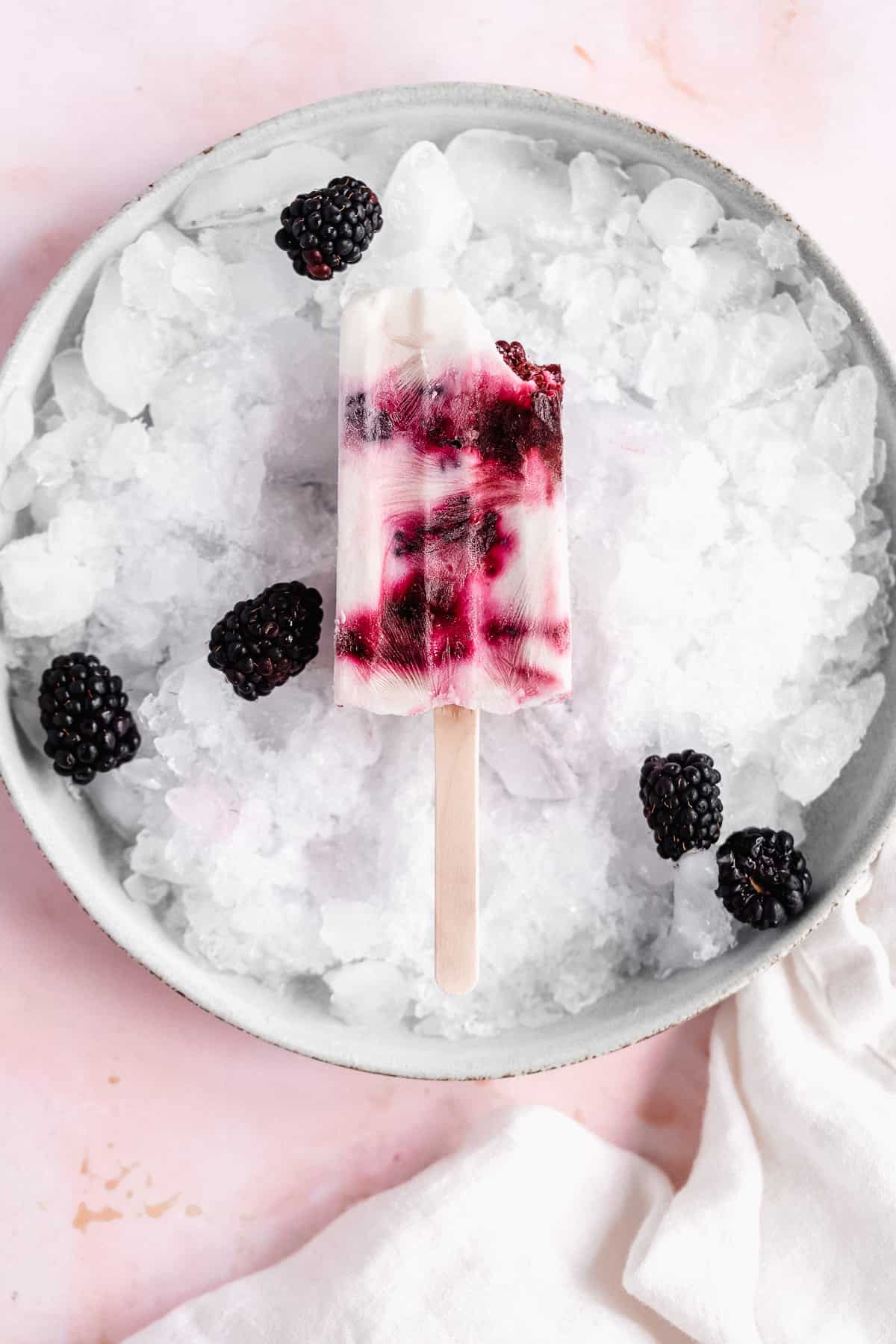 Overhead photo of a single popsicle sitting on top of ice on a plate.  A bite has been taken out of the popsicle.  Blackberries are sprinkled around.  
