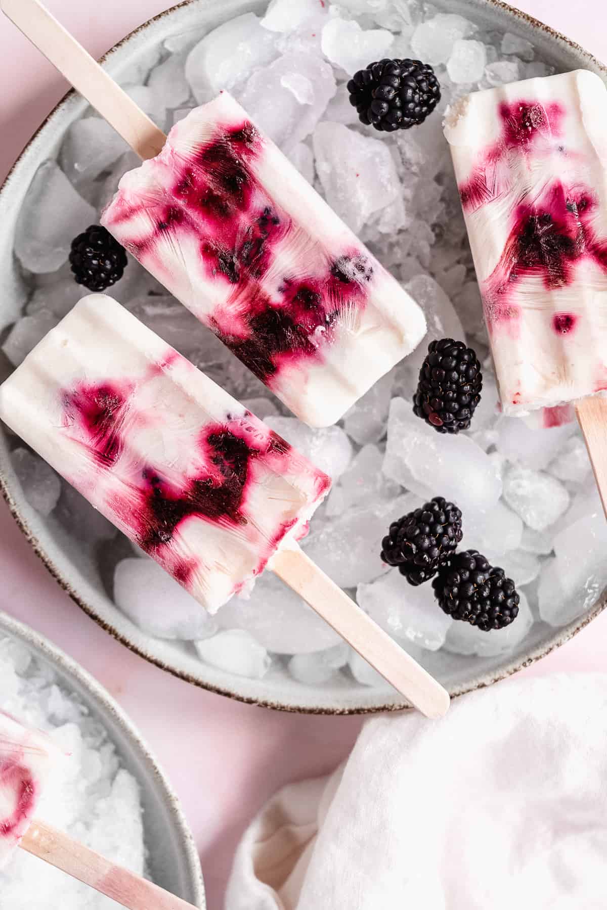Overhead photo of three blackberry popsicles on a plate with ice.  Blackberries are sprinkled around the popsicles.  