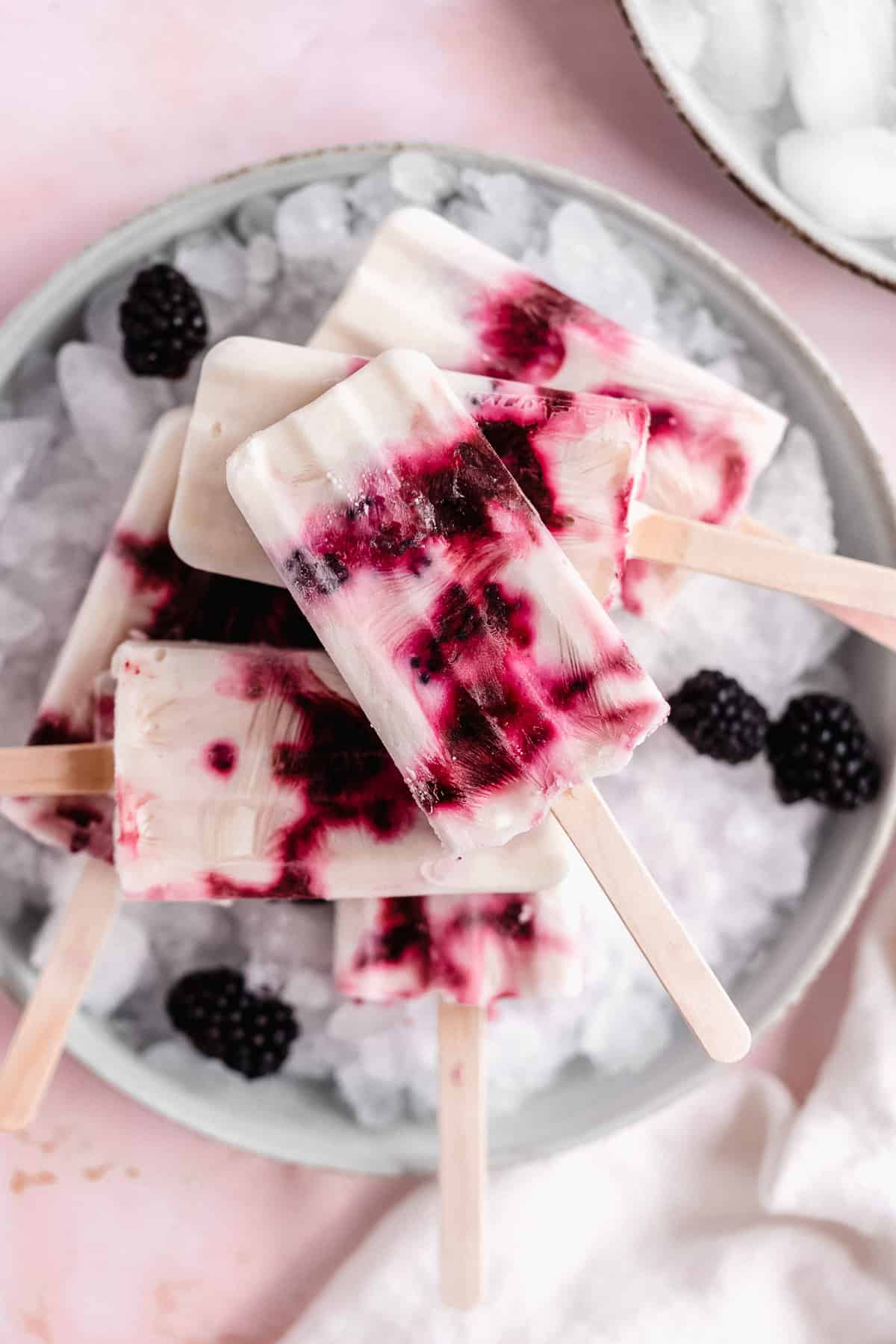 Overhead photo of a stack of blackberry popsicles on a layer of ice sitting on a white plate.  Extra blackberries are scattered around.   