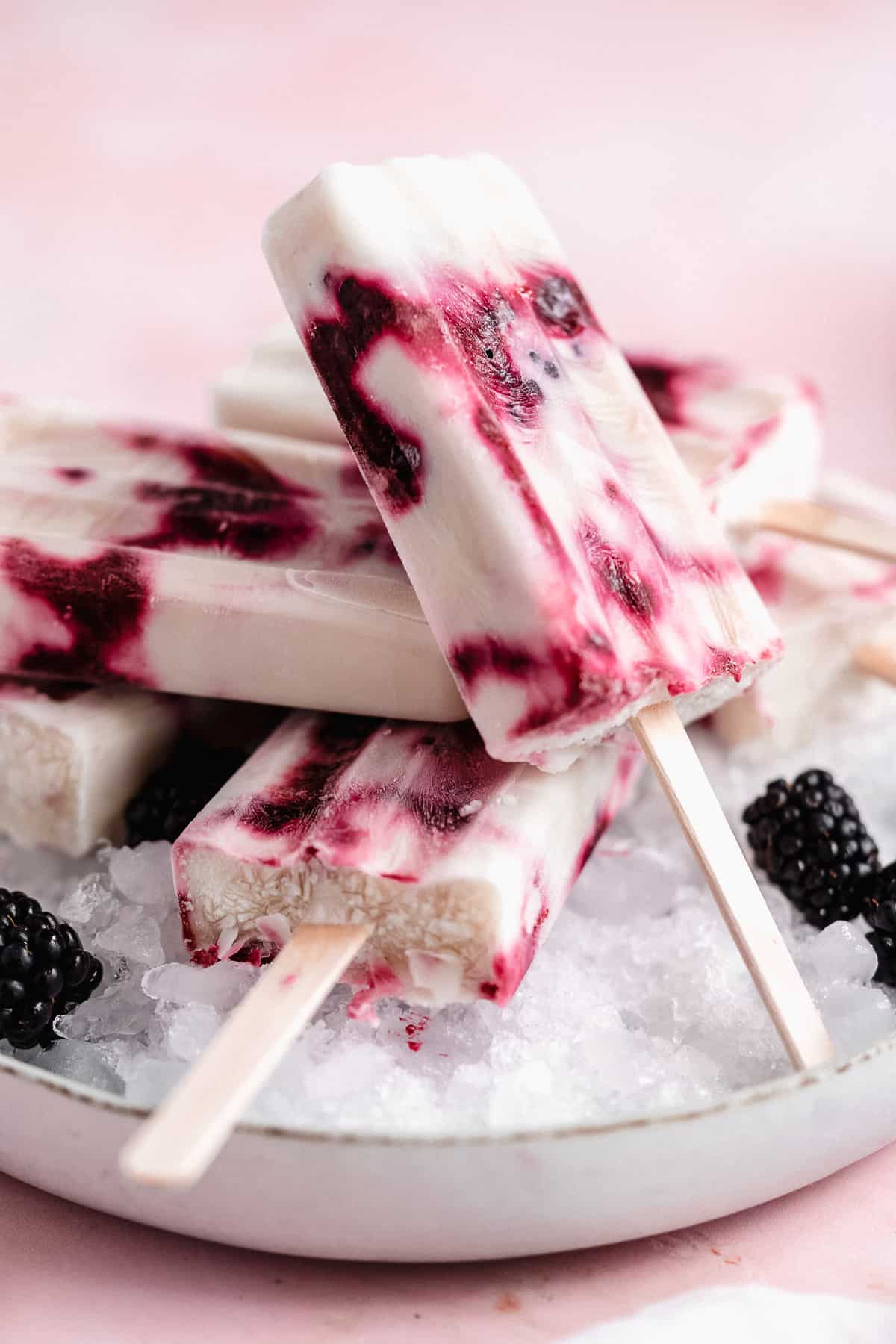 Side view closeup photo of several popsicles mounded on a layer of ice on a white plate.  Extra blackberries are sprinkled around.  