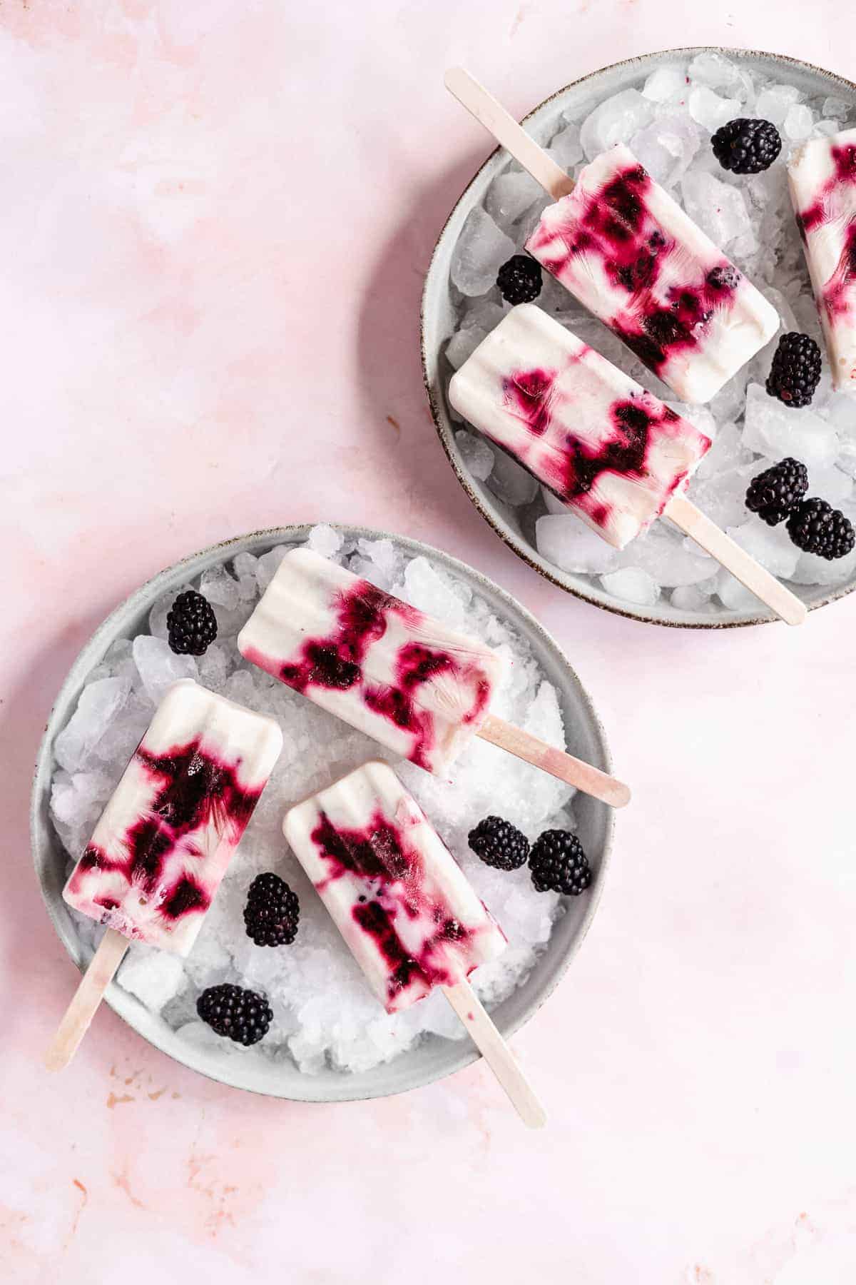 Overhead photo of two white plates - each with a layer of ice and three popsicles.  Extra blackberries are scattered around.  