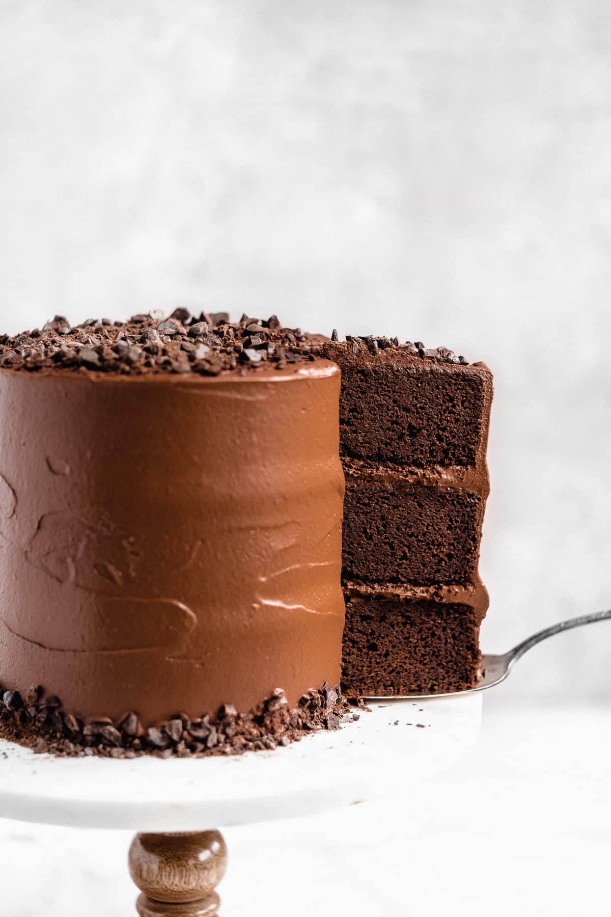 Side view closeup photo of the Blackout Chocolate Paleo Cake sitting on a white marble cake plate.  A piece of cake has been cut and is in the process of being removed with a cake server.  