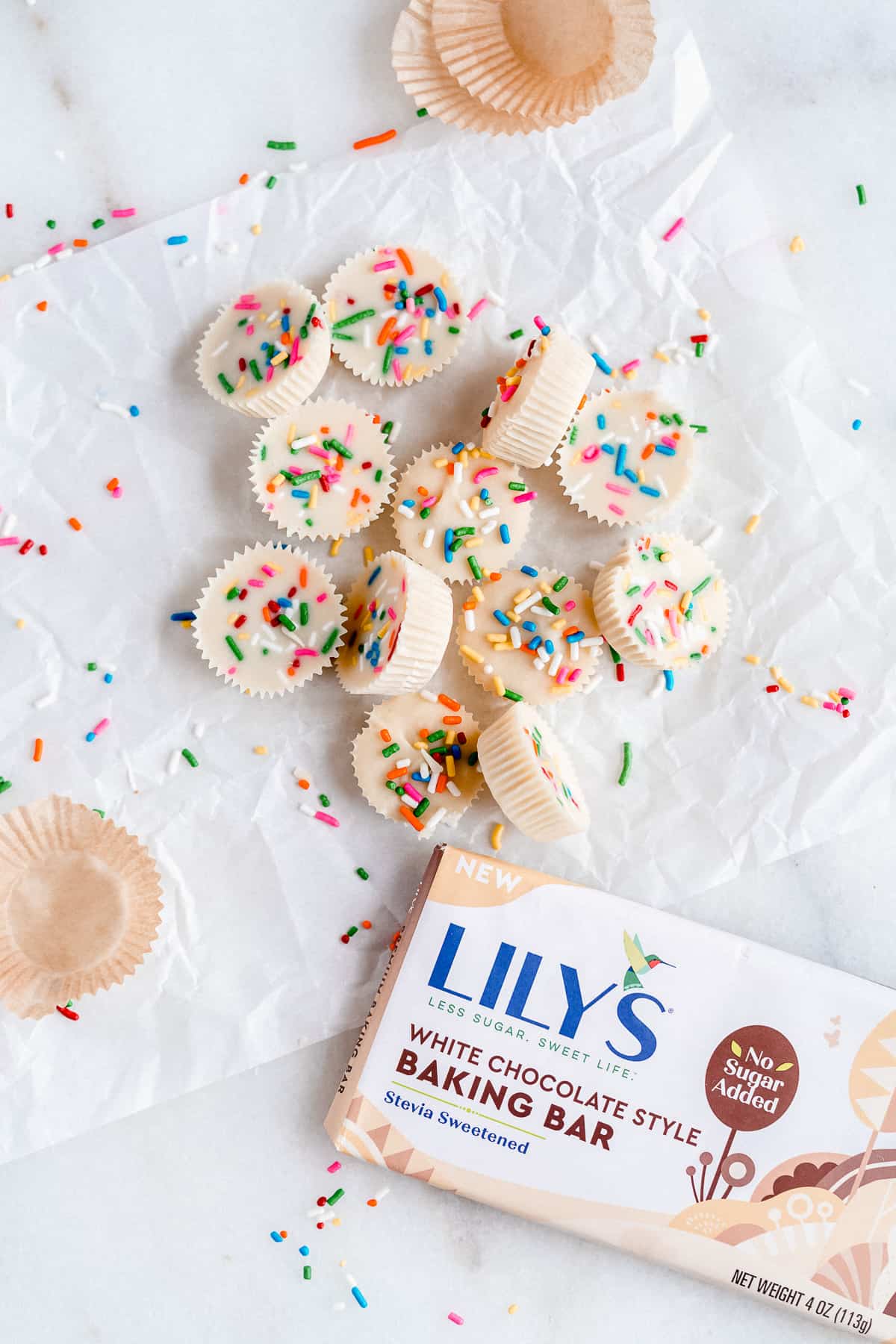 Pile of white chocolate peanut butter cups on white parchment paper with rainbow sprinkles and Lily's White Chocolate bar in the corner