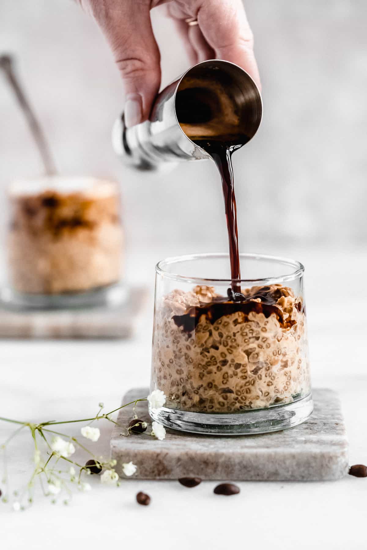 Side view photo of a small jar of Rich and Creamy Cappuccino Overnight Oats sitting on a marble coaster.  A hand is overhead pouring some extra espresso on top of the oats.  