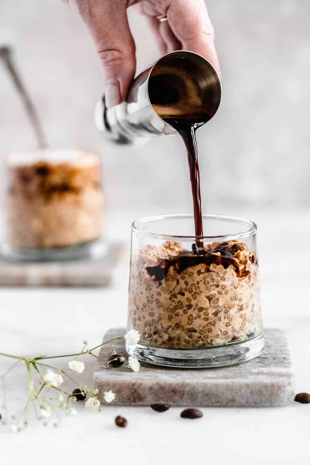 Side view photo of a jar of Rich and Creamy Cappuccino Overnight Oats sitting on a marble coaster. A hand is pouring some extra espresso on top of the oats. A second jar of oats can be seen in the background.