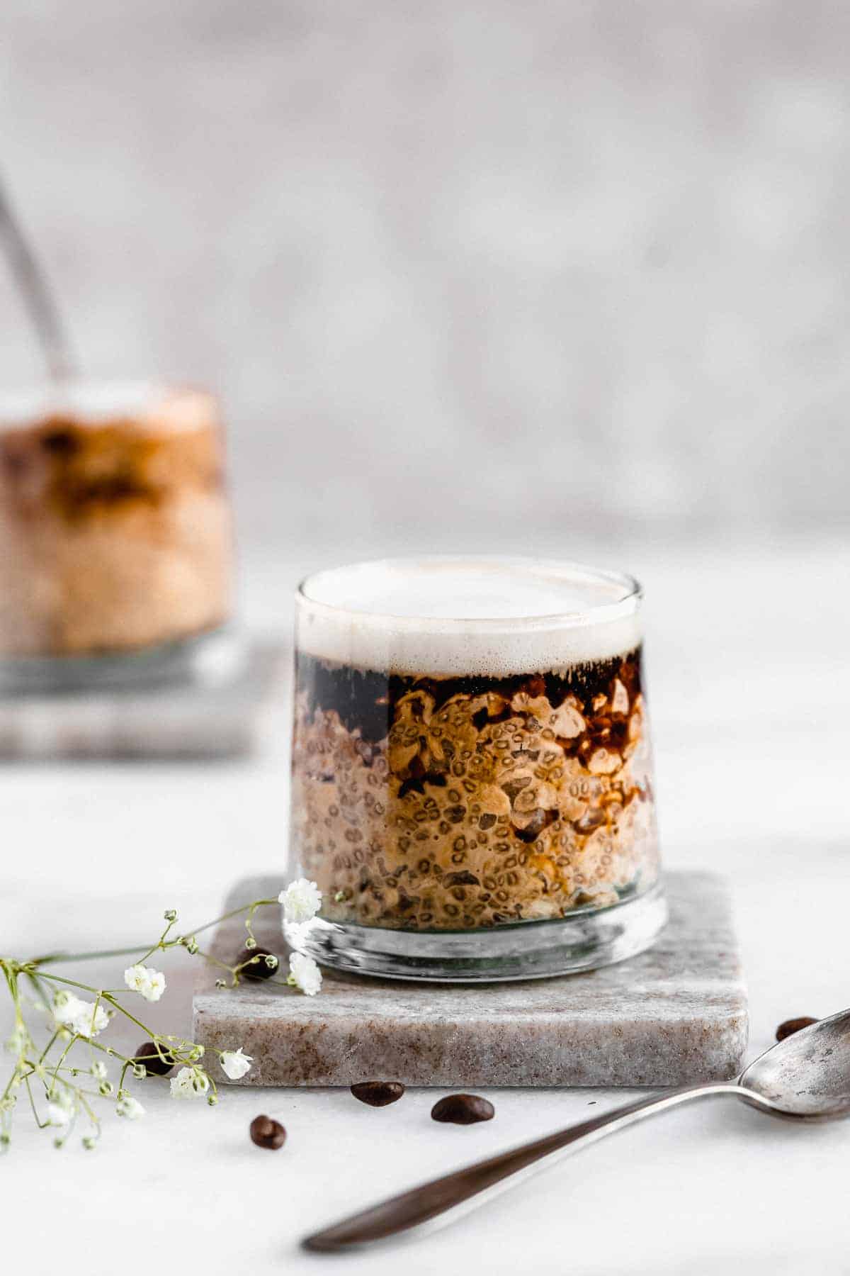 A side view photo of a jar of the completed Rich and Creamy Cappuccino Overnight Oats with a bit of espresso on top ready to be enjoyed. A silver spoon is laying nearby.