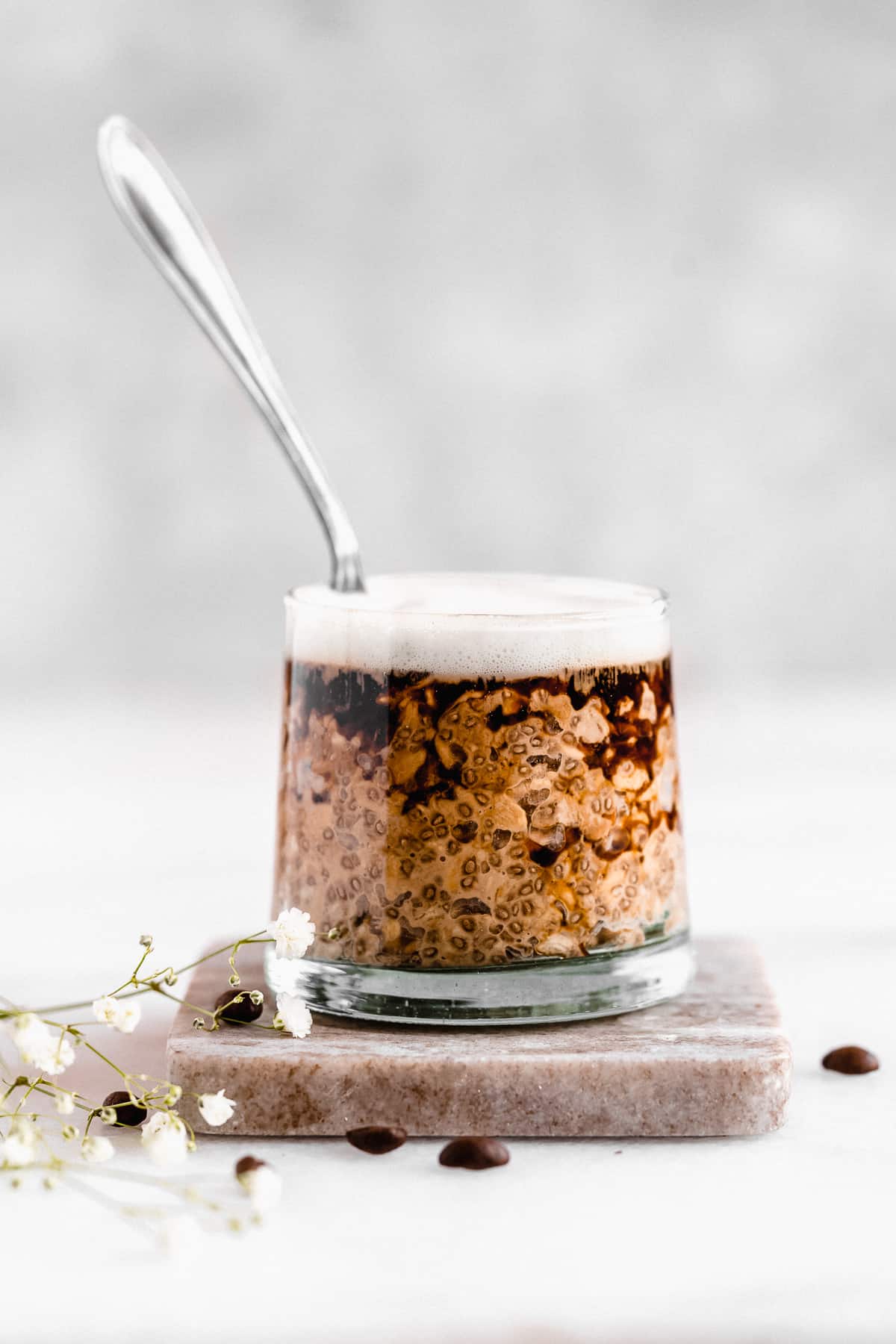 Side view photo of a jar of Rich and Creamy Cappuccino Overnight Oats topped with a bit of espresso sitting on a marble coaster.  A silver spoon is sticking up in the middle of the oats.  