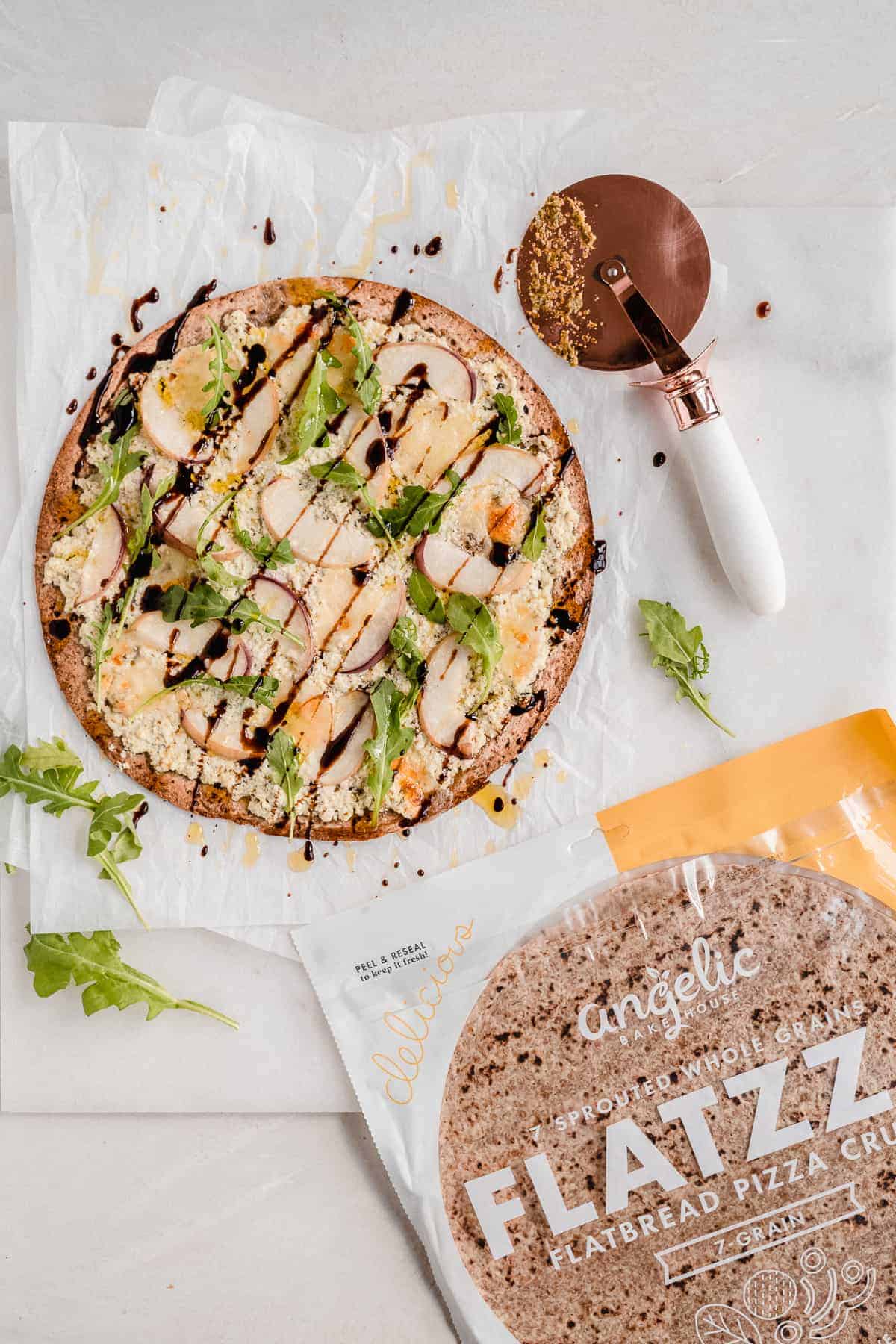 Overhead photo of a whole Crispy White Peach and Ricotta Pesto Flatbread with Balsamic Glaze sitting on white parchment paper on a marble slab.  A pizza cutter and a bag of Angelic Bakehouse Flatbread Pizza Crusts are laying nearby.  