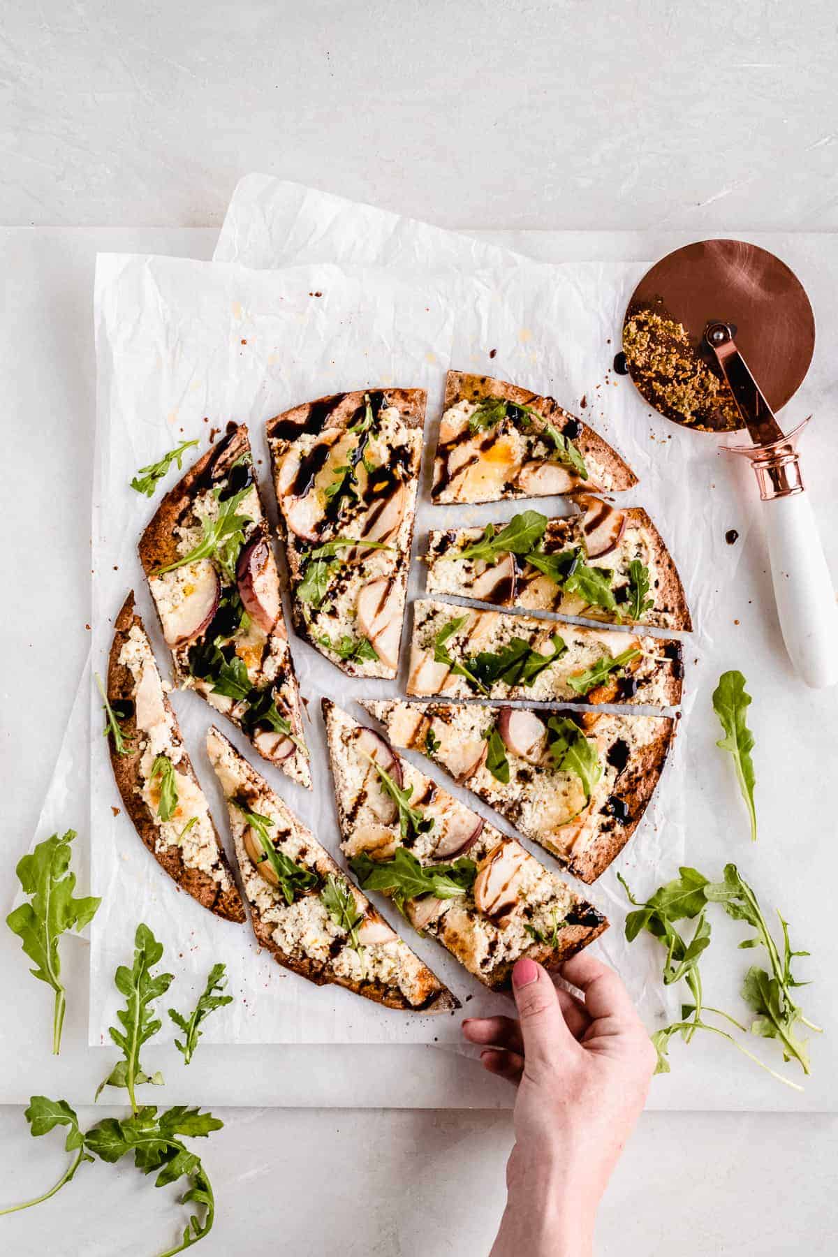 Overhead photo of a Crispy White Peach and Ricotta Pesto Flatbread with Balsamic Glaze placed on top of white parchment paper on a marble slab that has been cut into slices that are separated. A pizza cutter is laying nearby. A hand is pulling one piece away.