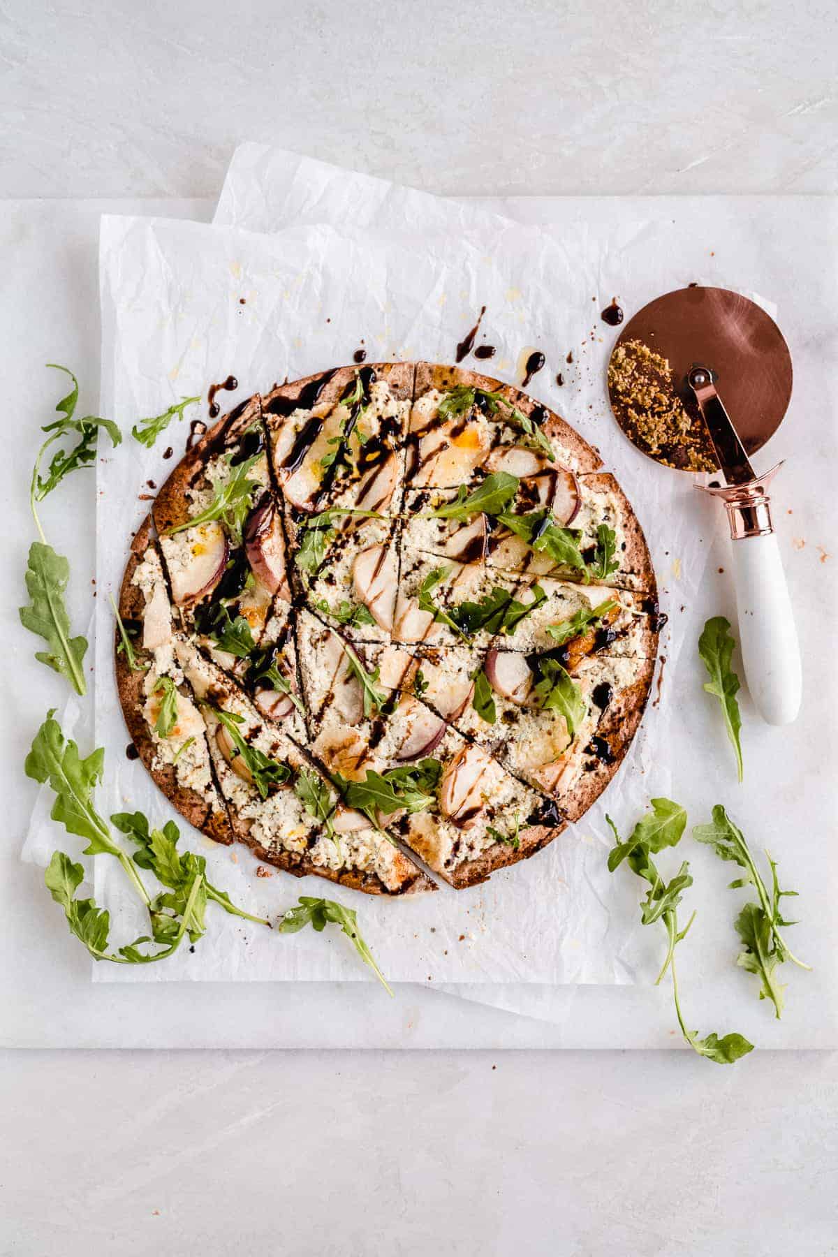 Overhead photo of the Crispy White Peach and Ricotta Pesto Flatbread with Balsamic Glaze laying on white parchment paper on a white marble slab that has been cut into pieces.  A pizza cutter is laying nearby.  