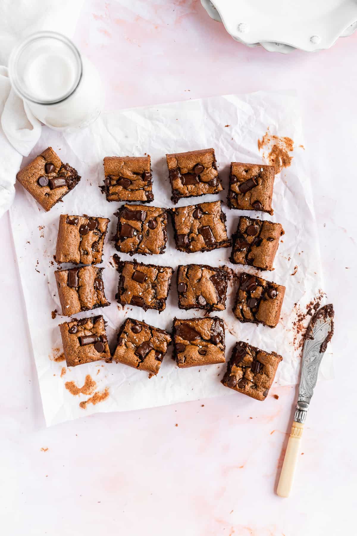 Overhead photo of the completed recipe of Gluten-free Almond Butter Blondies cut into 16 square pieces and arranged on white parchment paper.  The silver knife that cut the pieces is laying nearby.  