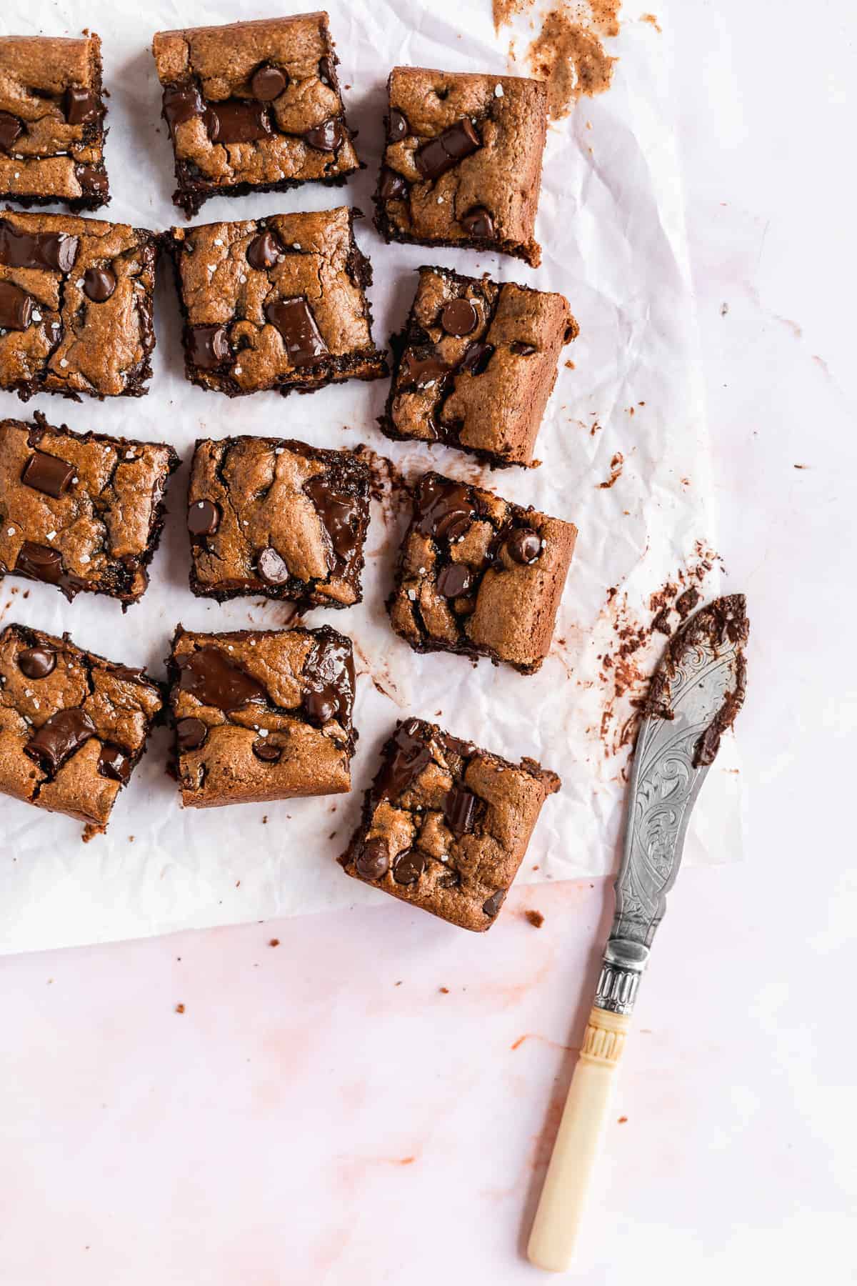 Overhead photo of the completed recipe of Gluten-free Almond Butter Blondies cut into squares and separated on white parchment paper. A silver knife is laying nearby.