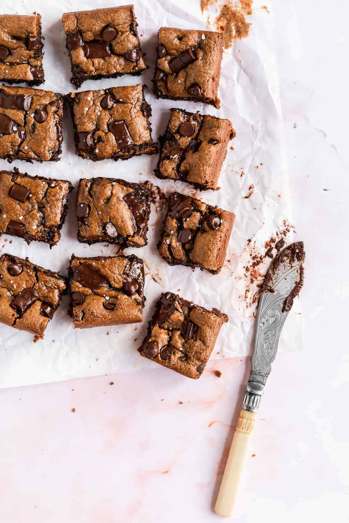 Overhead photo of the completed recipe of Gluten-free Almond Butter Blondies cut into squares and separated on white parchment paper.  A silver knife is laying nearby.  