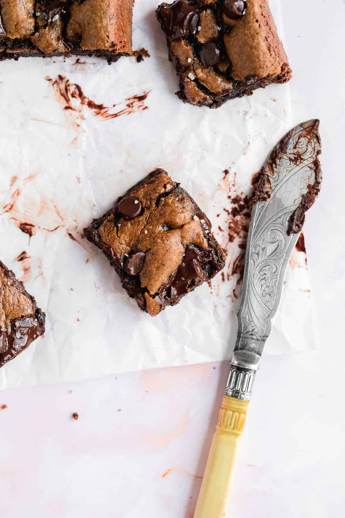 Overhead photo of several Gluten-free Almond Butter Blondies cut into square pieces and arranged on white parchment paper.  A silver knife that cut the pieces is sitting nearby.  