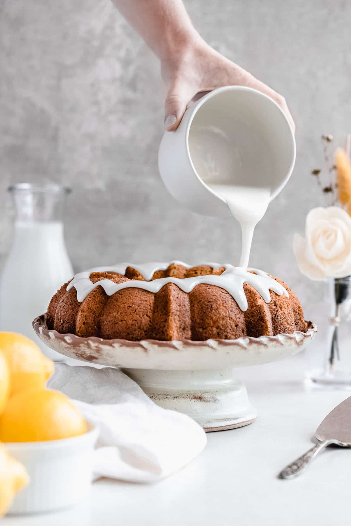 Side view photo of the freshly baked Citrus-y Gluten Free Lemon Poppy Seed Bundt Cake sitting on a cake plate with a hand holding a jar of icing from above and pouring the icing over the cake.  A carafe of milk and a bowl of lemons can be seen in the background.  