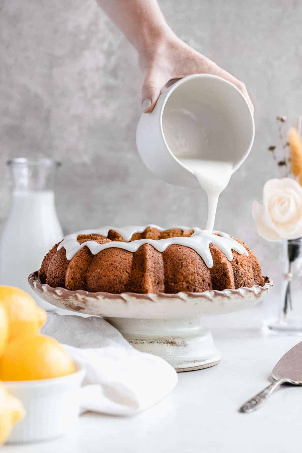 Side view photo of the freshly baked Citrus-y Gluten Free Lemon Poppy Seed Bundt Cake sitting on a cake plate with a hand holding a jar of icing from above and pouring the icing over the cake. A carafe of milk and a bowl of lemons can be seen in the background.