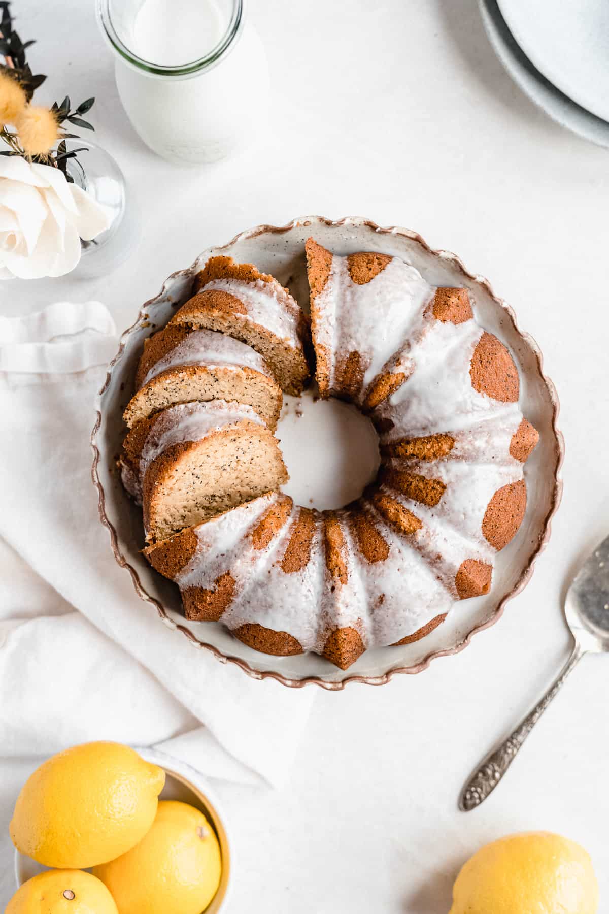 Overhead photo of the freshly baked and iced Citrus-y Gluten Free Lemon Poppy Seed Bundt Cake sitting on a cake plate with scalloped edges.  Several slices have been cut.  A silver cake server is laying nearby.  A bowl of lemons and carafe of milk can be seen in the background.  