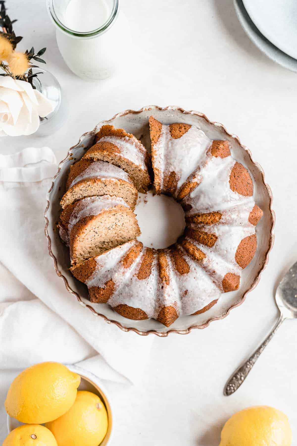 Overhead photo of the freshly baked and iced Citrus-y Gluten Free Lemon Poppy Seed Bundt Cake sitting on a cake plate with scalloped edges. Several slices have been cut. A silver cake server is laying nearby. A bowl of lemons and carafe of milk can be seen in the background.