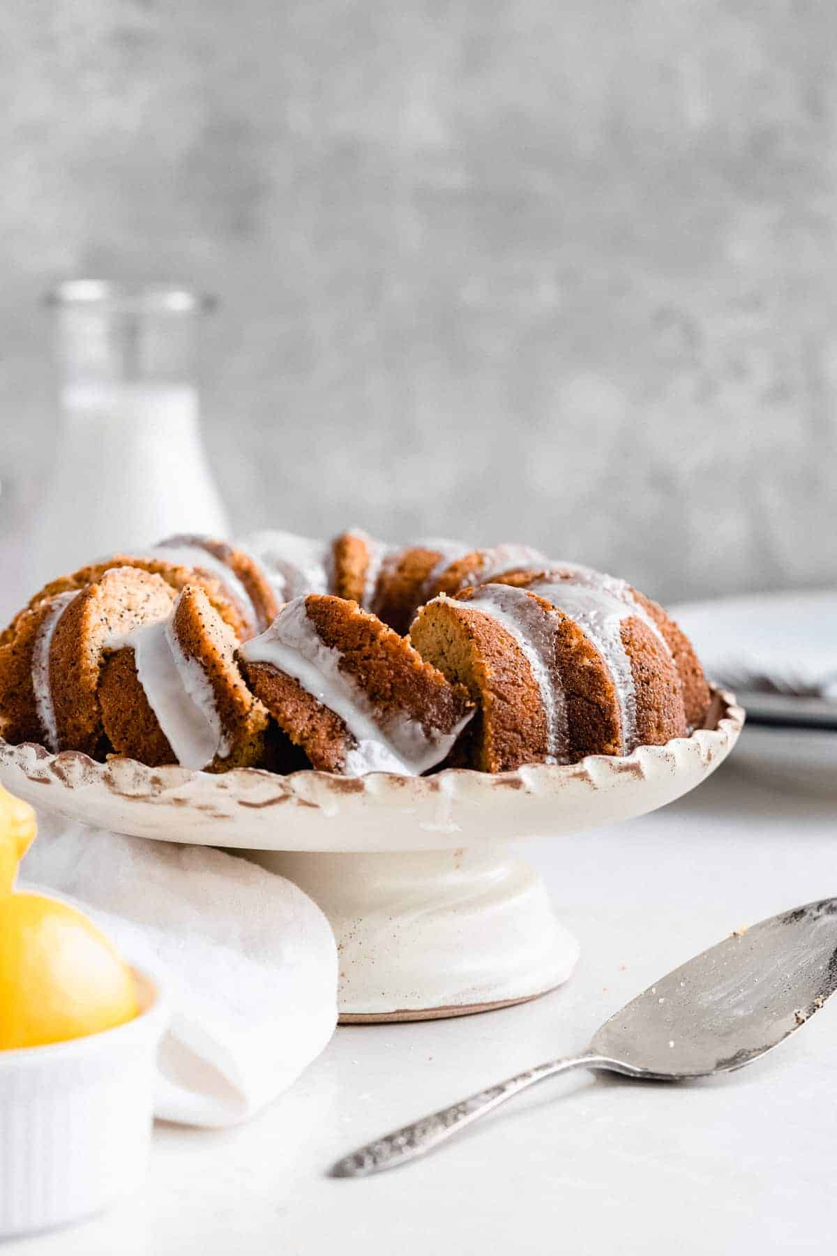 Side view photo of the freshly baked and iced Citrus-y Gluten Free Lemon Poppy Seed Bundt Cake sitting on a cake plate with scalloped edges.  Several pieces of cake are sliced and a cake server is laying nearby.  A carafe of milk and bowl of lemons can be seen in the background.  