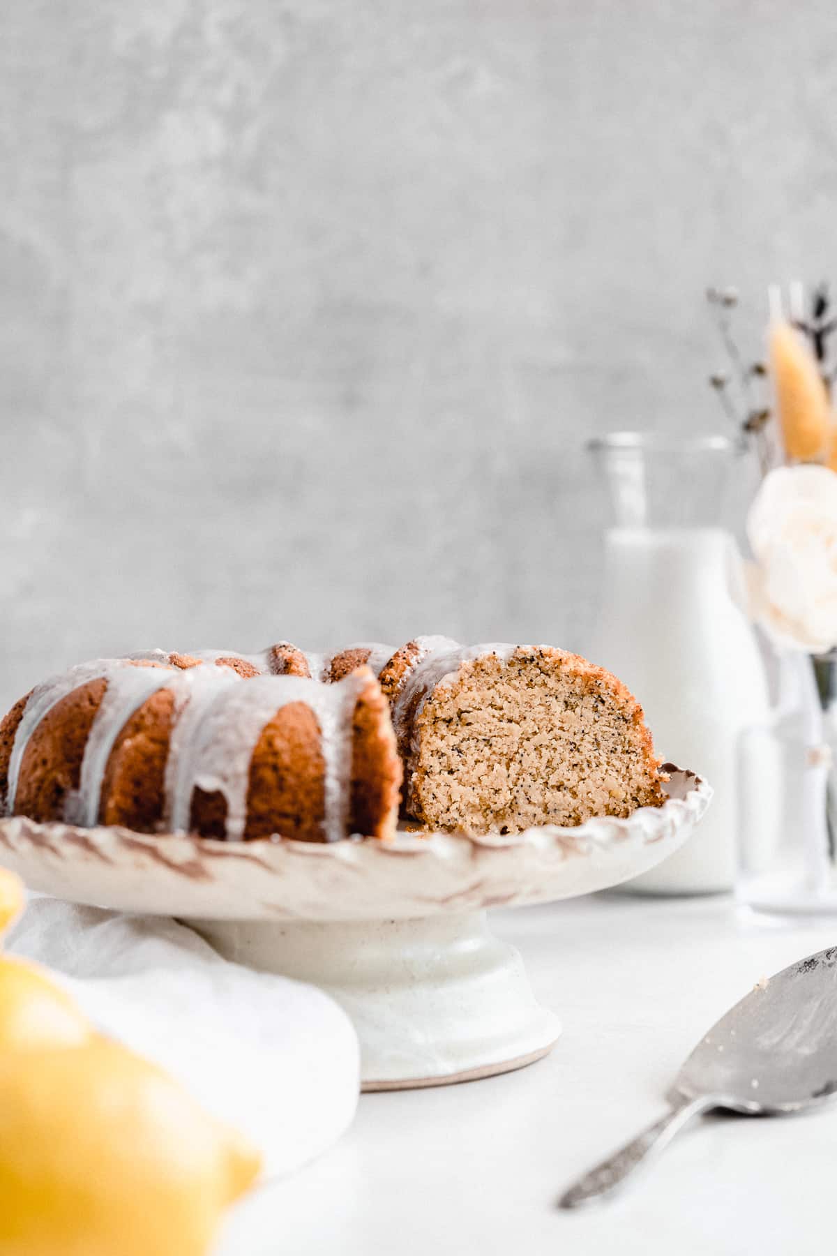 Side view closeup of the Citrus-y Gluten Free Lemon Poppy Seed Bundt Cake sitting on a cake plate with scalloped edges. Several pieces have been cut and removed. A silver cake server is laying nearby and a carafe of milk and bowl of lemons can be seen in the background.