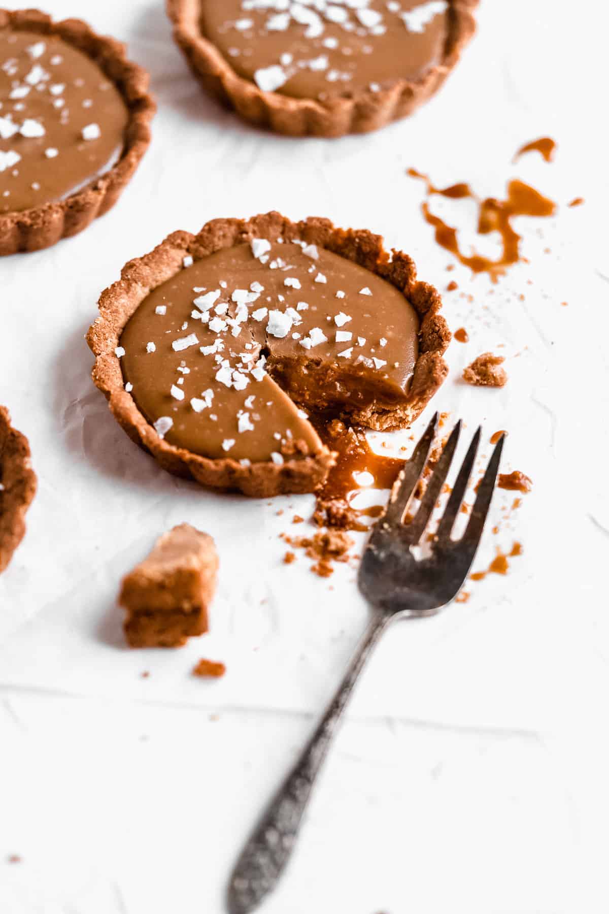 Side view closeup of several Vegan Salted Caramel Tarts arranged on white parchment paper.  One tart has a bite missing and a fork laying nearby.  