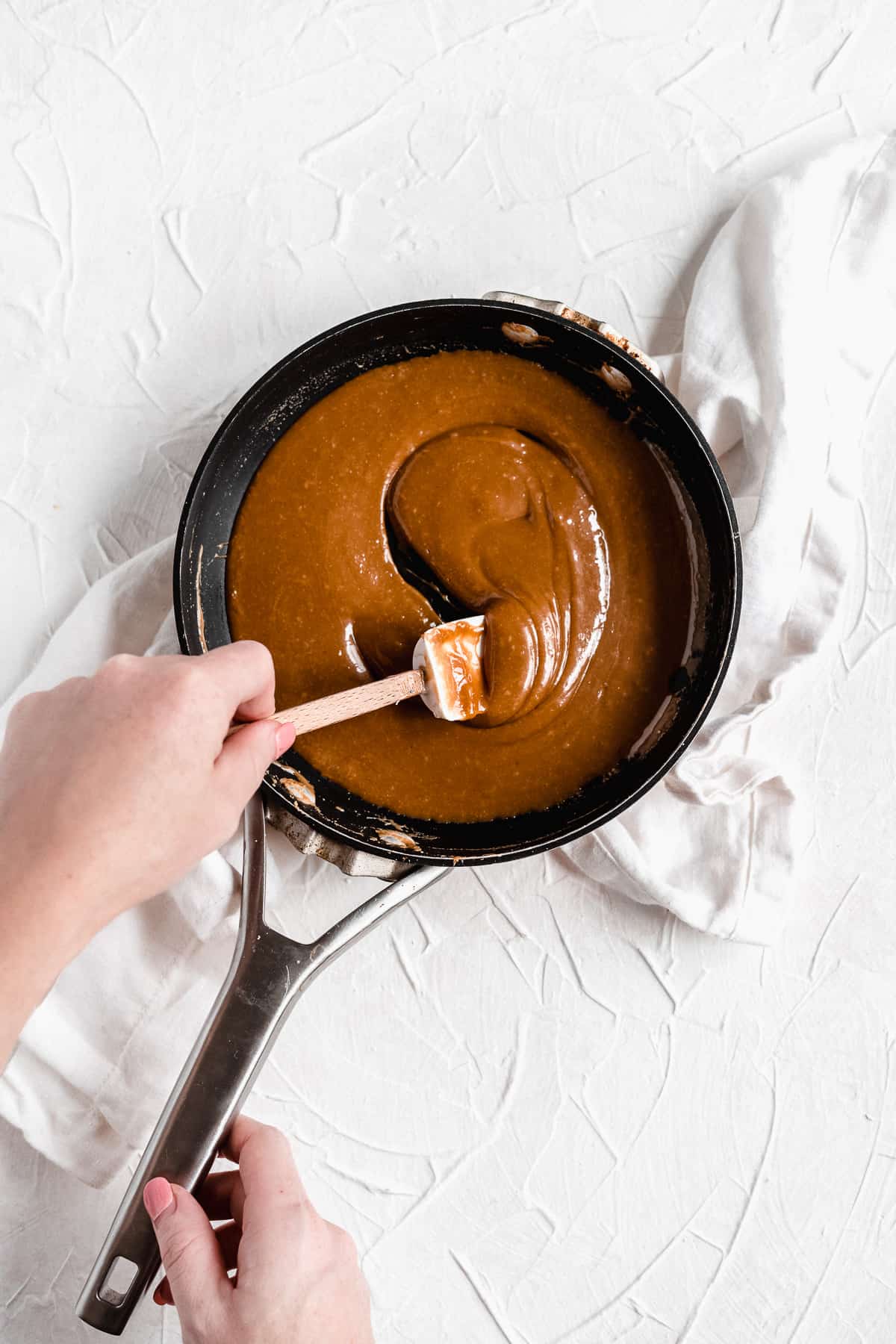 An overhead photo of the salted caramel mixture being made in a black saucepan.  A hand with a white spatula is stirring the mixture.  