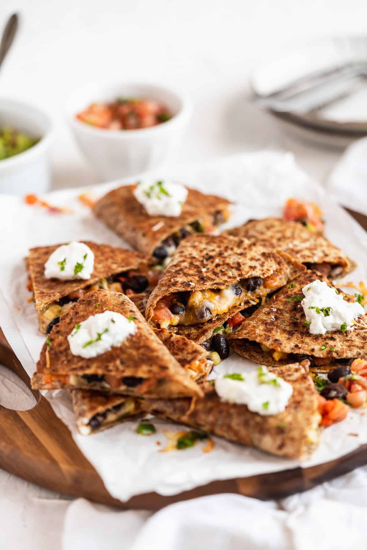 Overhead photo of Southwest Quesadilla triangles ready to be enjoyed.  They are mounded on parchment paper on a wooden tray.  Several of the triangles are topped with a dollop of greek yogurt.  