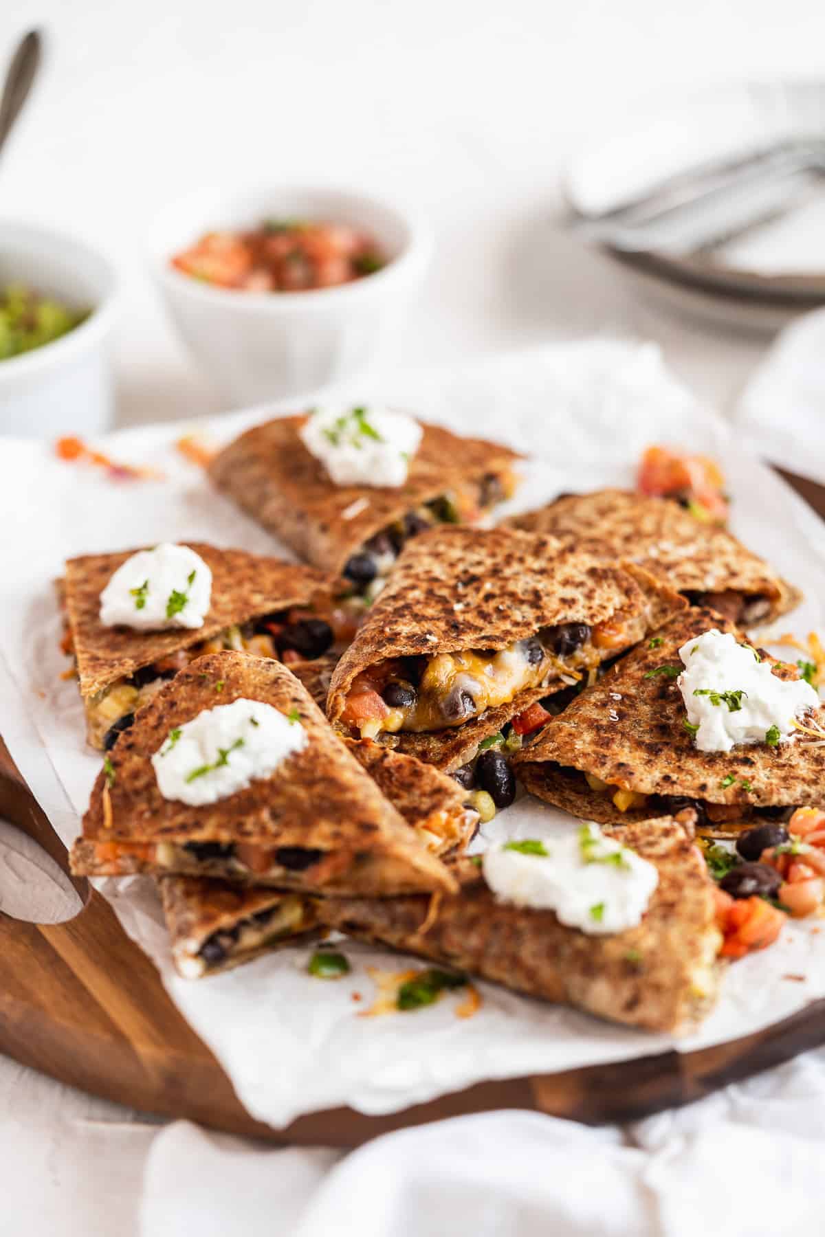 Overhead photo of Southwest Quesadilla triangles ready to be enjoyed. They are mounded on parchment paper on a wooden tray. Several of the triangles are topped with a dollop of greek yogurt.