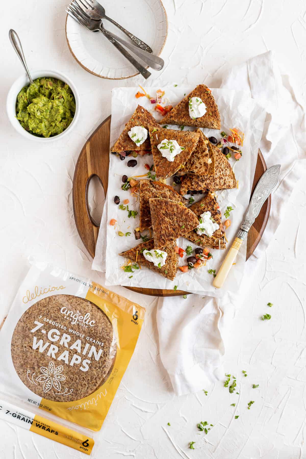 Overhead photo of Southwest Quesadilla triangles stacked on parchment paper on a wooden tray.  A bowl of guacamole and small plates with forks are sitting nearby.  A bag of Angelic Bakehouse 7 Grain Wraps is also nearby.  