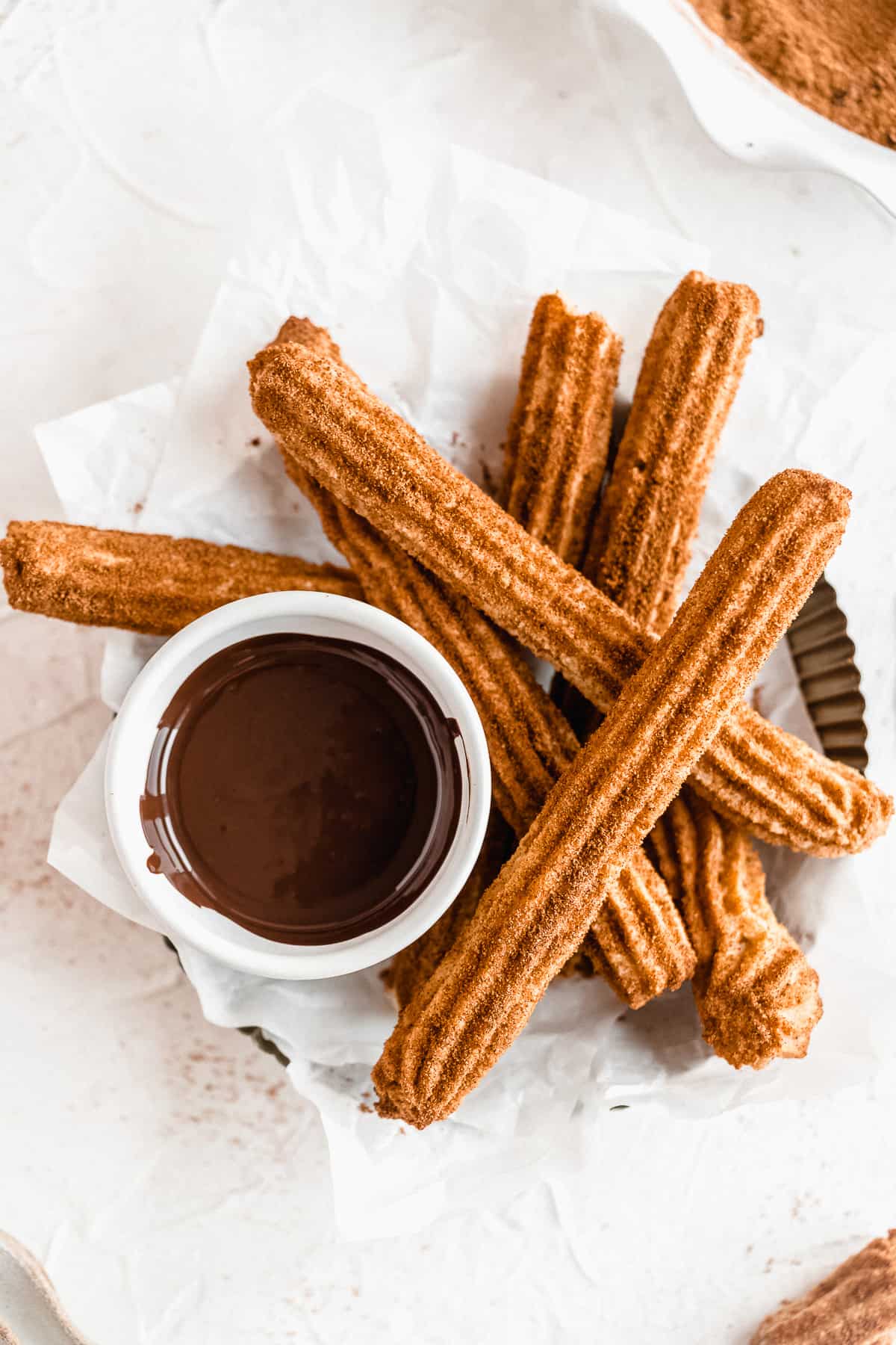 Basket of churros on a white surface with a bowl of melted chocolate.