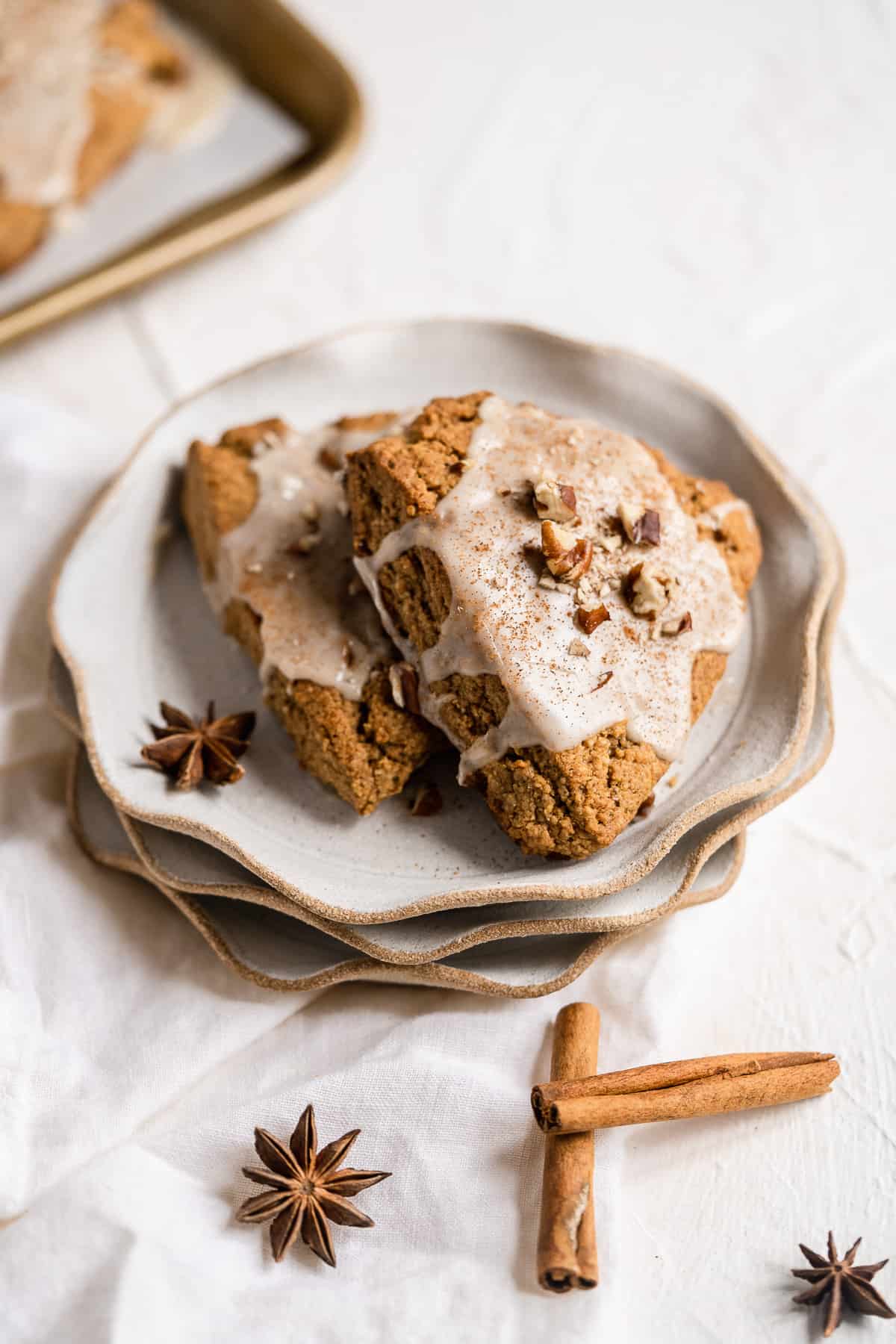 Close up overhead photo of two freshly baked and glazed Chai Spiced Gluten-free Vegan Scones sitting on a stack of three small scalloped plates. Cinnamon sticks are laying nearby.