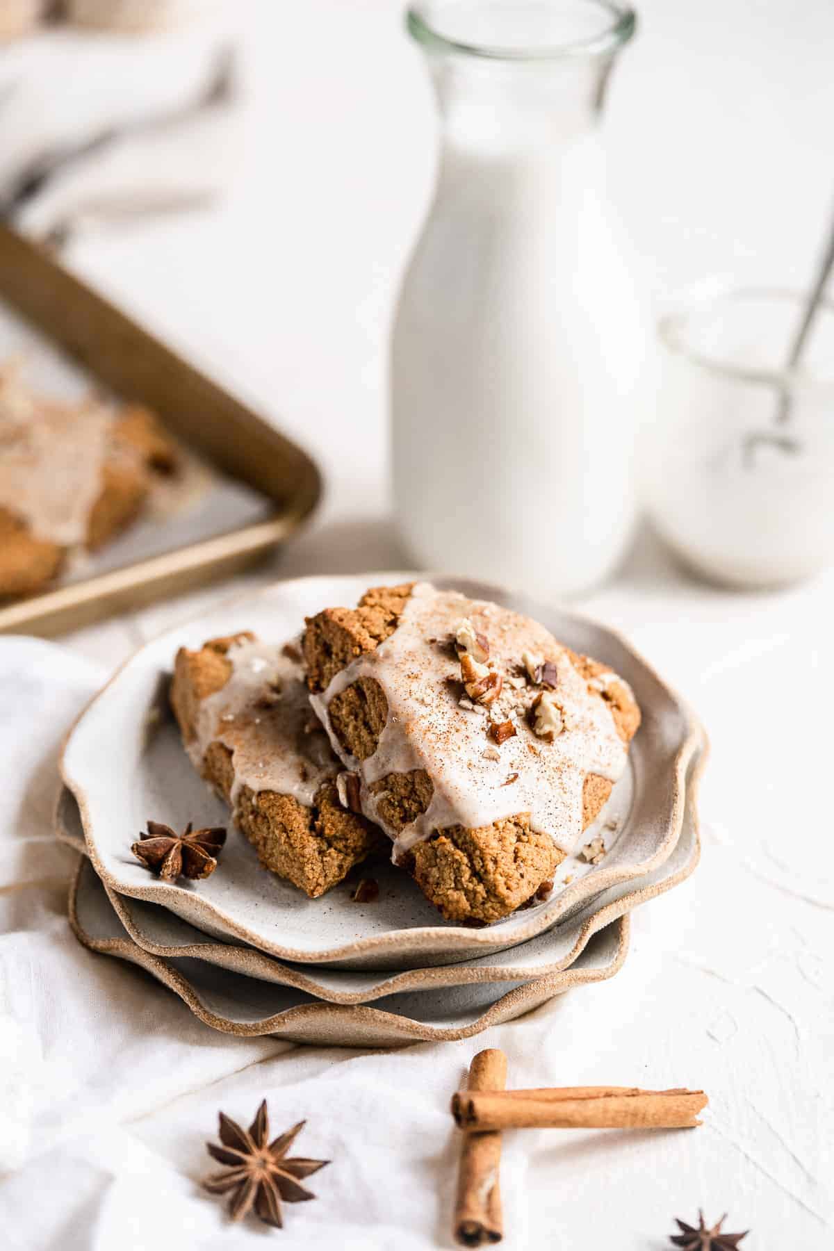 Close up view of two Chai Spiced Gluten-free Vegan Scones sitting on a stack of three small plates.  A glass of milk and a jar of glaze are in the background.  