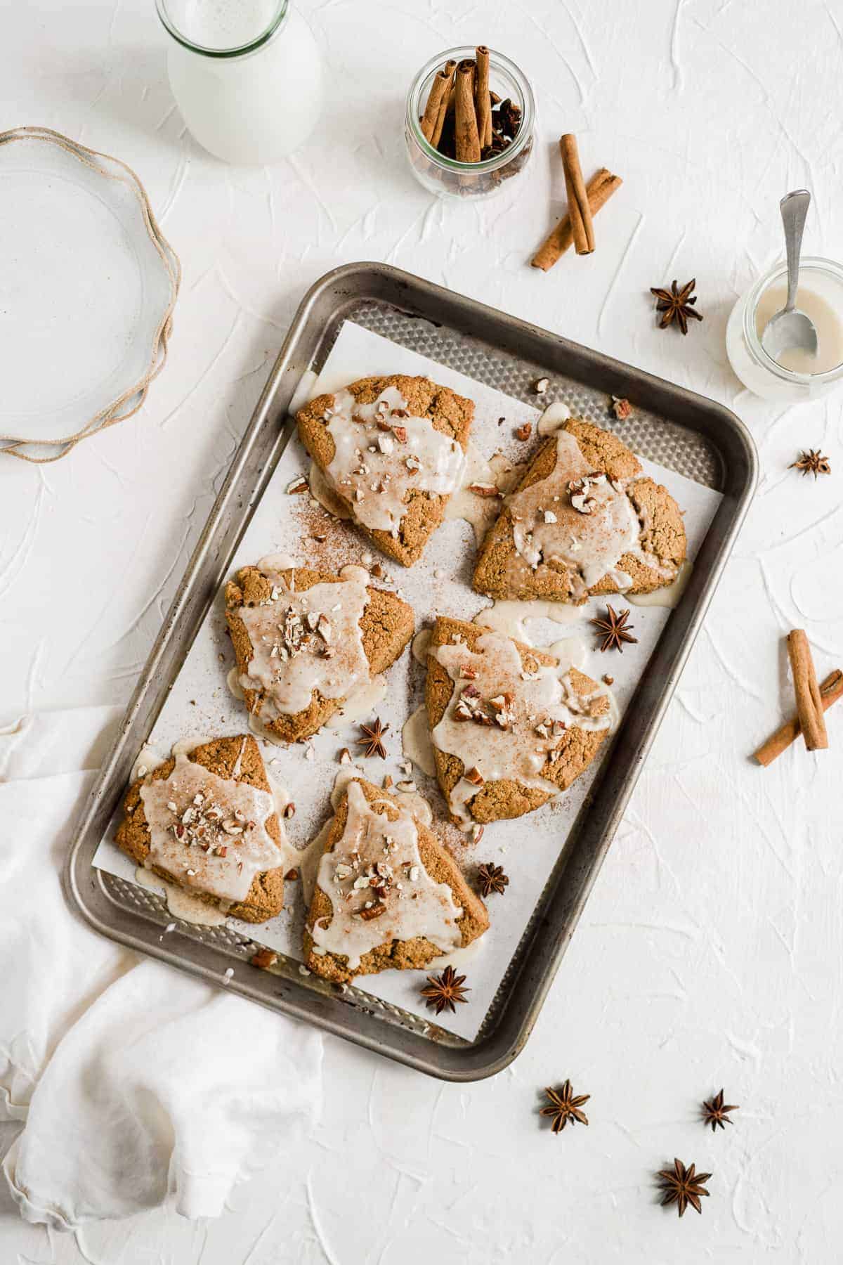 Overhead photo of a baking sheet with parchment paper and six freshly baked and glazed Chai Spiced Gluten-free Vegan Scones. Some milk, a jar of glaze and several small plates are sitting nearby.
