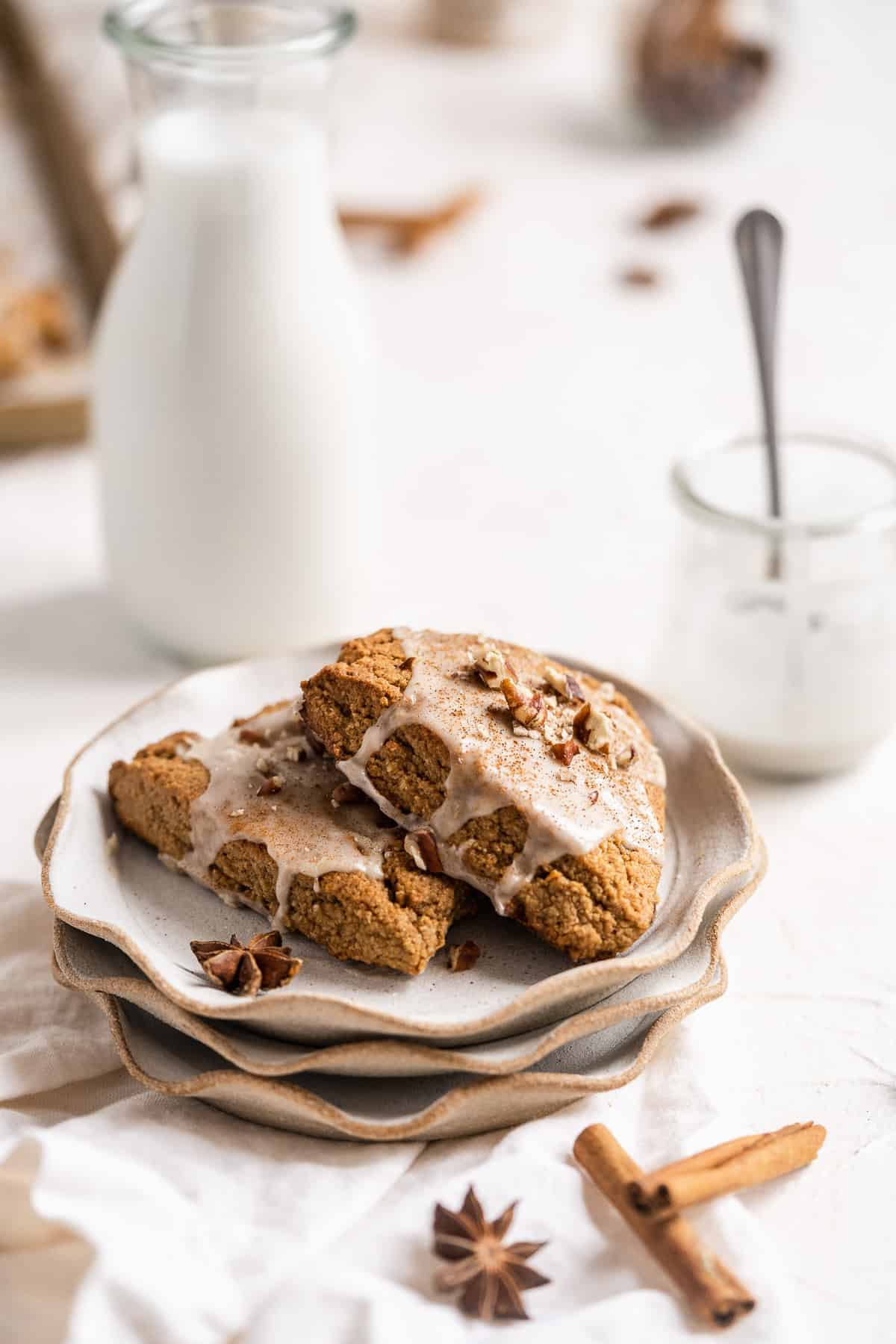 Side view close up of two Chai Spiced Gluten-free Vegan Scones sitting on three stacked small plates.  A glass of milk and jar of glaze are in the background.  