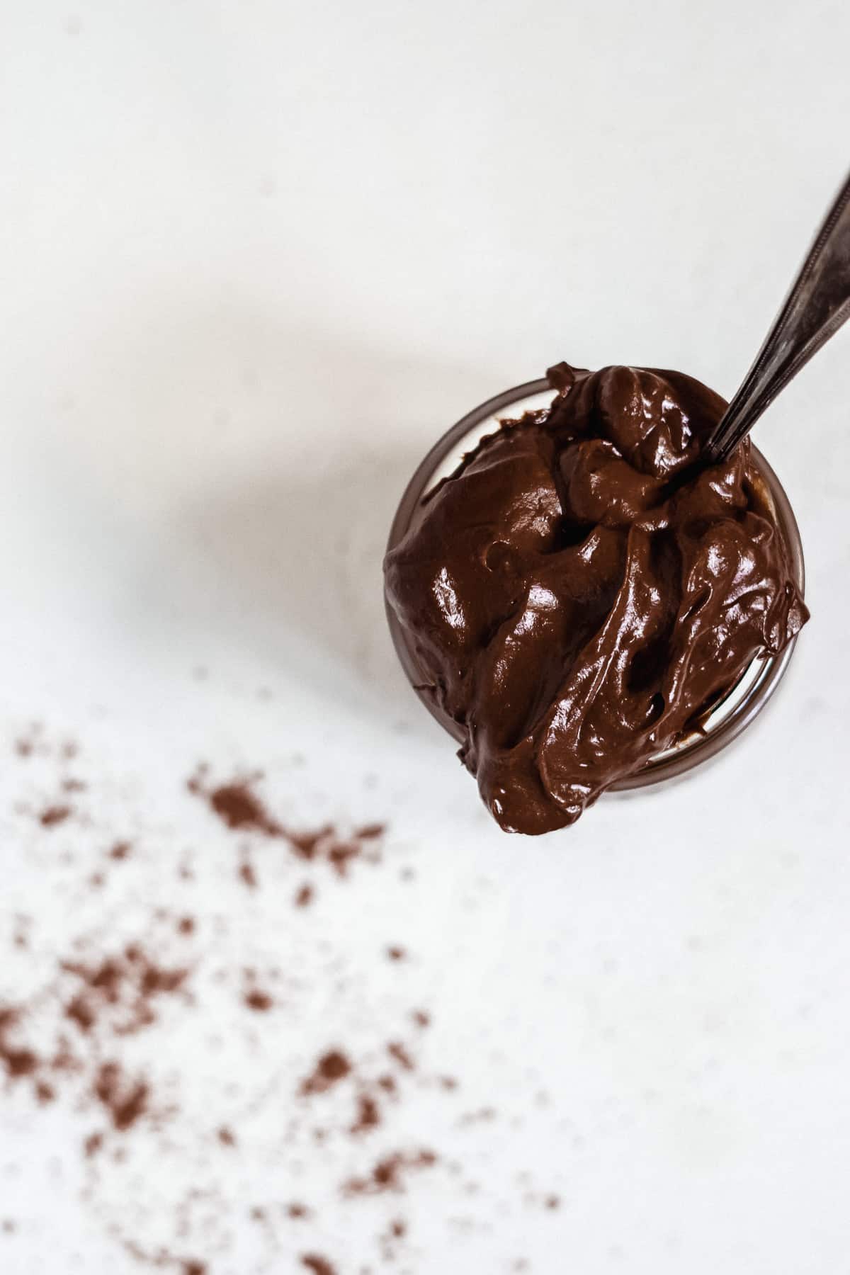 Overhead view of a jar with chocolate frosting on a white surface.