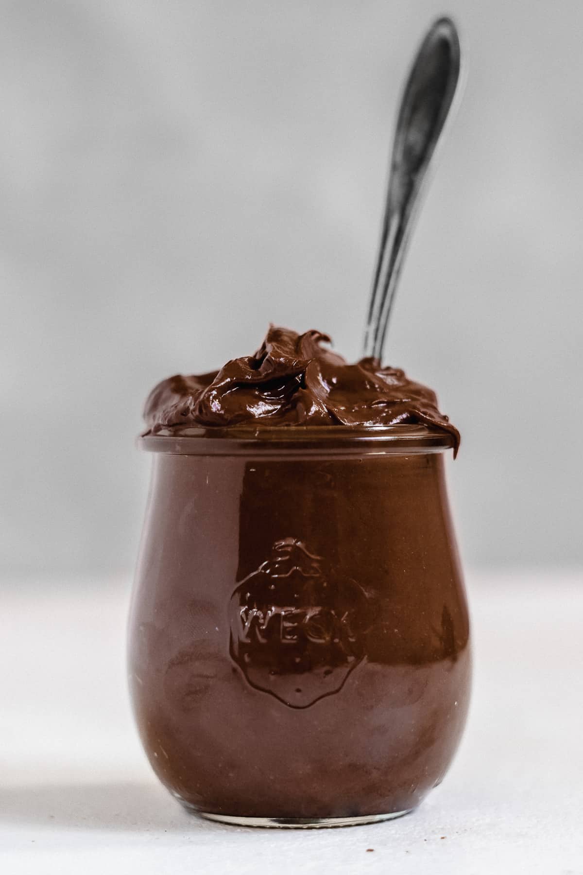 Jar overflowing with chocolate frosting with a spoon on a grey background.