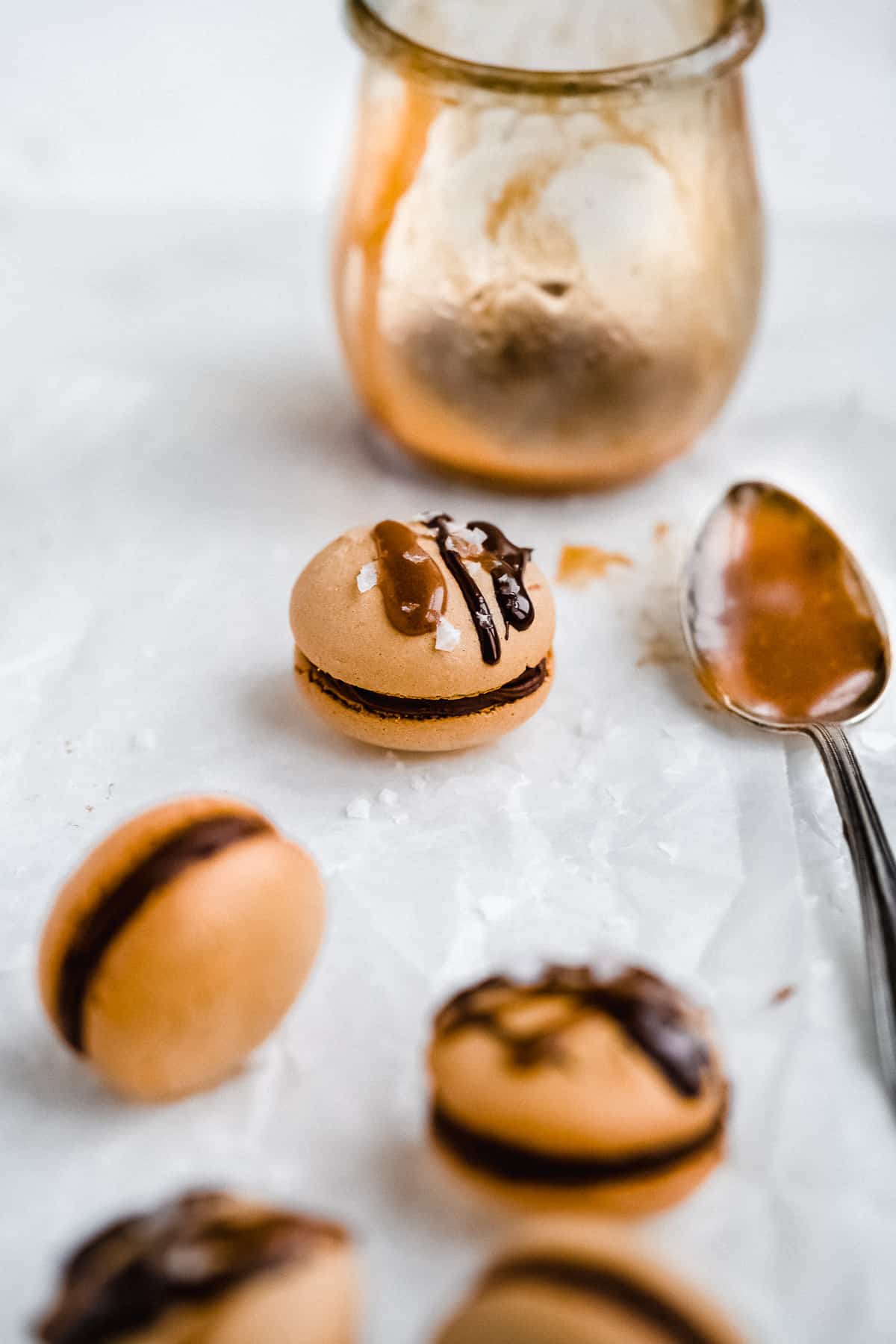 Close up photo of several Dark Chocolate Caramel Dairy-free Macarons on parchment paper on a white marble slab.  A jar with the caramel sauce and a spoon filled with caramel sauce are placed nearby.  