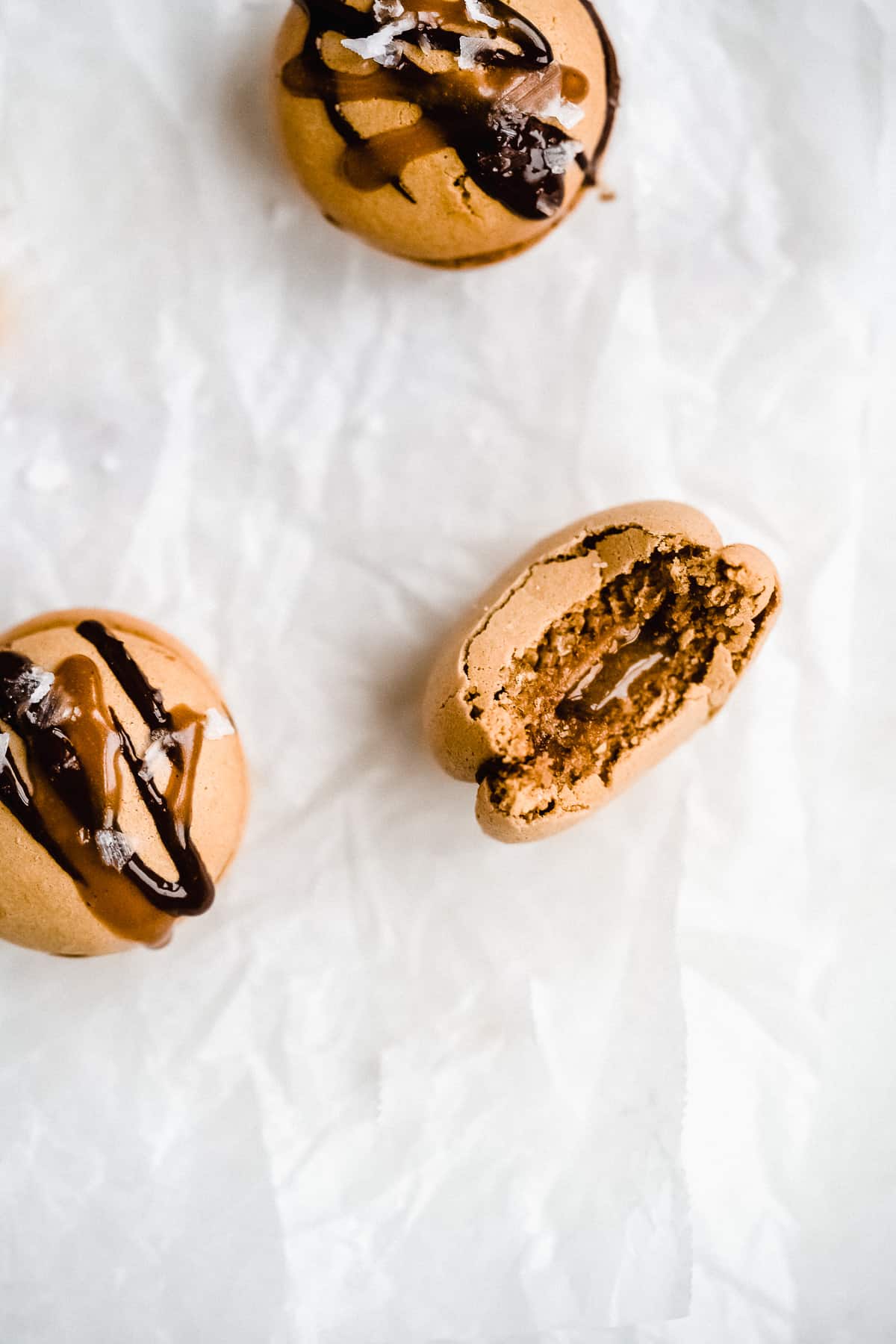 Close up photo of several Dark Chocolate Caramel Dairy-free Macarons sitting on parchment paper on a white marble slab.  One macaron has a bite taken out of it to reveal the delicious inside.  