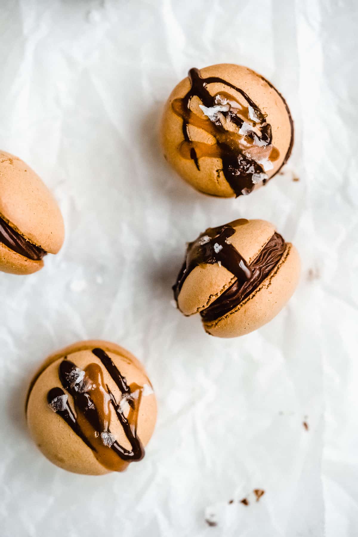 Overhead photo of several Dark Chocolate Caramel Dairy-free Macarons on white parchment paper and a marble slab.  One is laying on its side to show the filling inside.  