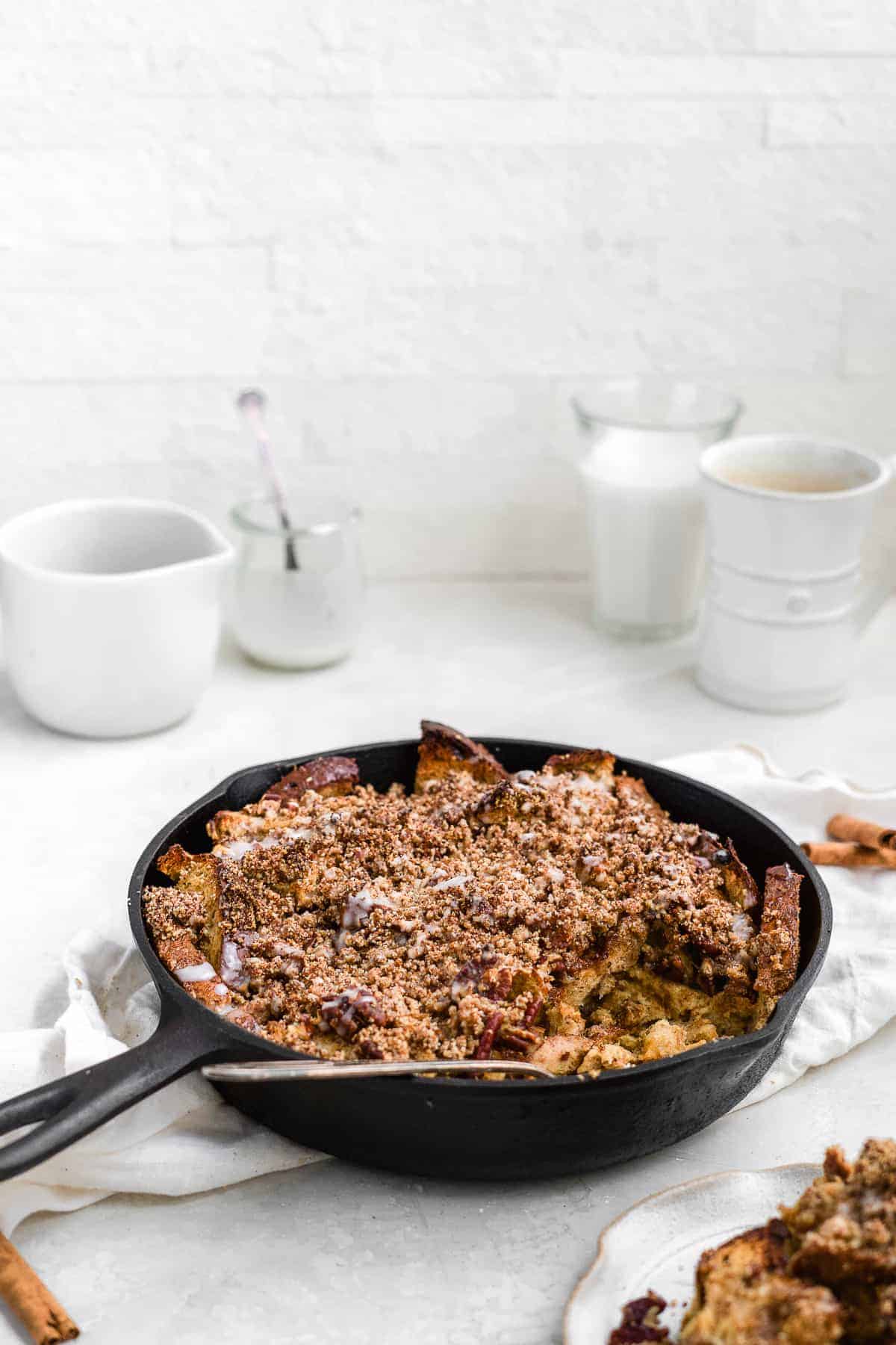 Warm and ready to be enjoyed this is an overhead photo of the Maple Cinnamon Raisin Dairy-free French Toast Skillet Casserole in a cast iron skillet sitting on a white dish towel on a marble slab. A serving of the casserole has been placed on a plate nearby. A glass of milk and a cup of coffee can be seen in the background.