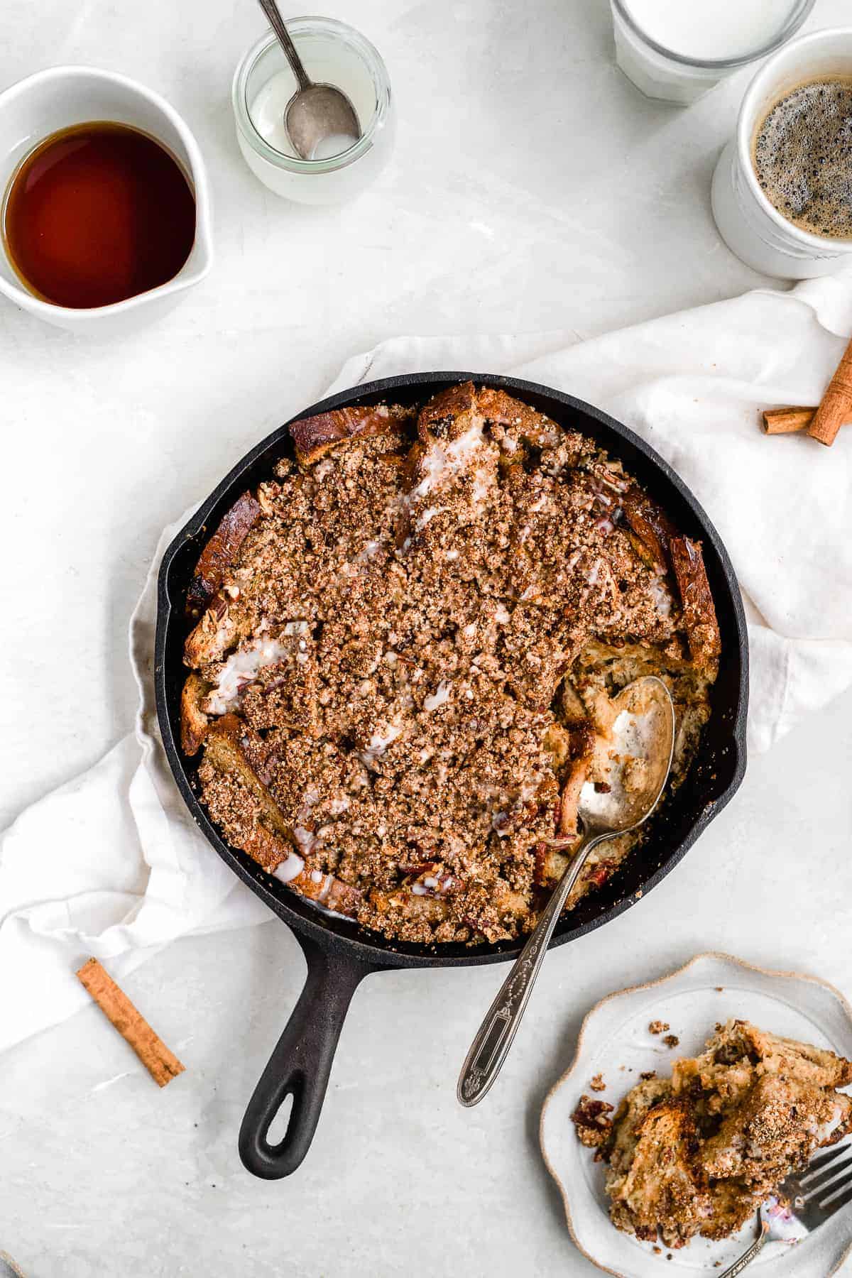 Overhead photo of a freshly baked Maple Cinnamon Raisin Dairy-free French Toast Skillet Casserole in a cast iron skillet with a serving spoon. A serving of the casserole has been placed on an adjacent white scalloped plate with a fork placed nearby. A glass of milk and cup of coffee can be seen in the background.