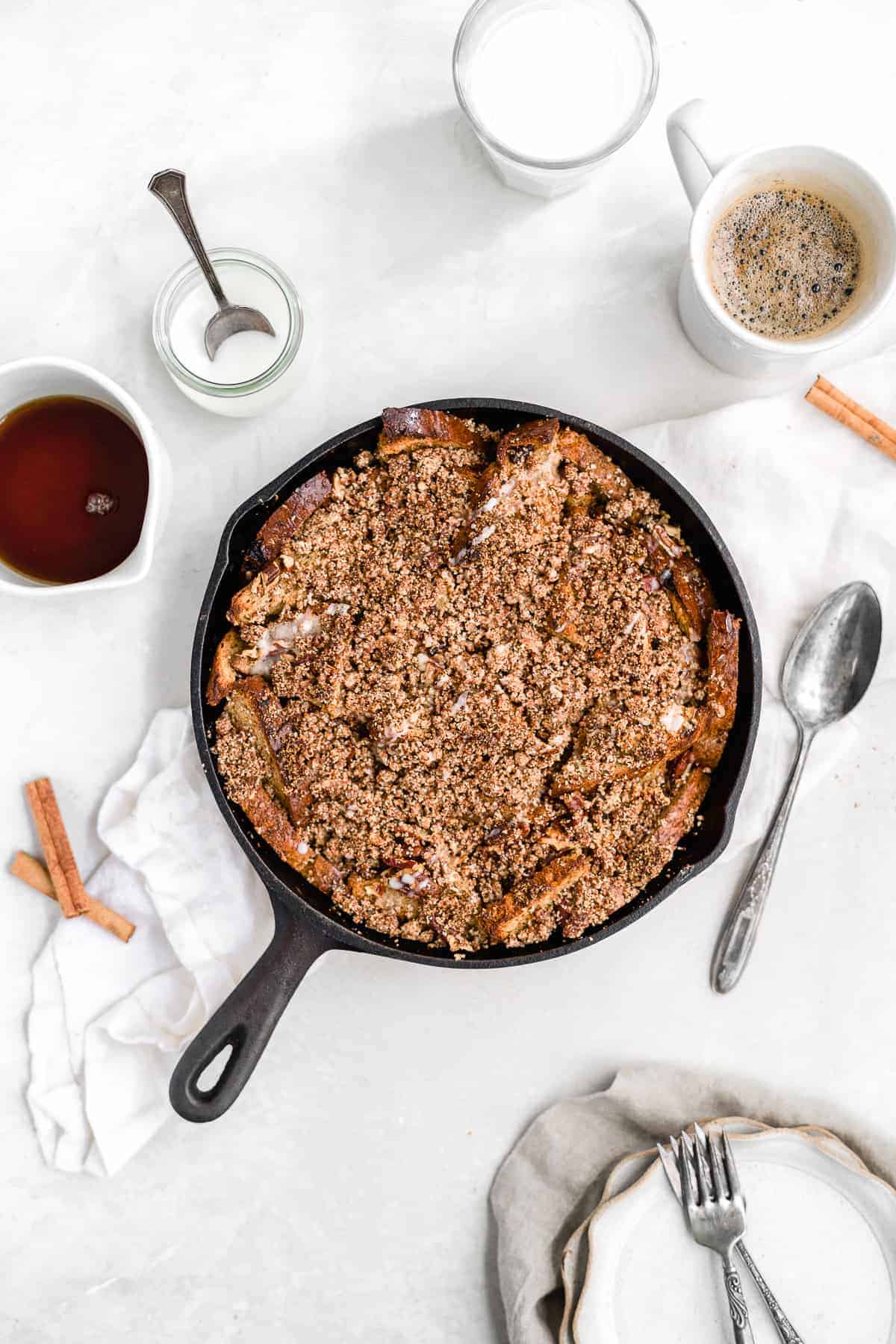 Overhead photo of the freshly baked Maple Cinnamon Raisin Dairy-free French Toast Skillet Casserole in a cast iron skillet on a white marble slab. A spoon, cinnamon sticks and several scalloped plates and forks are sitting nearby.