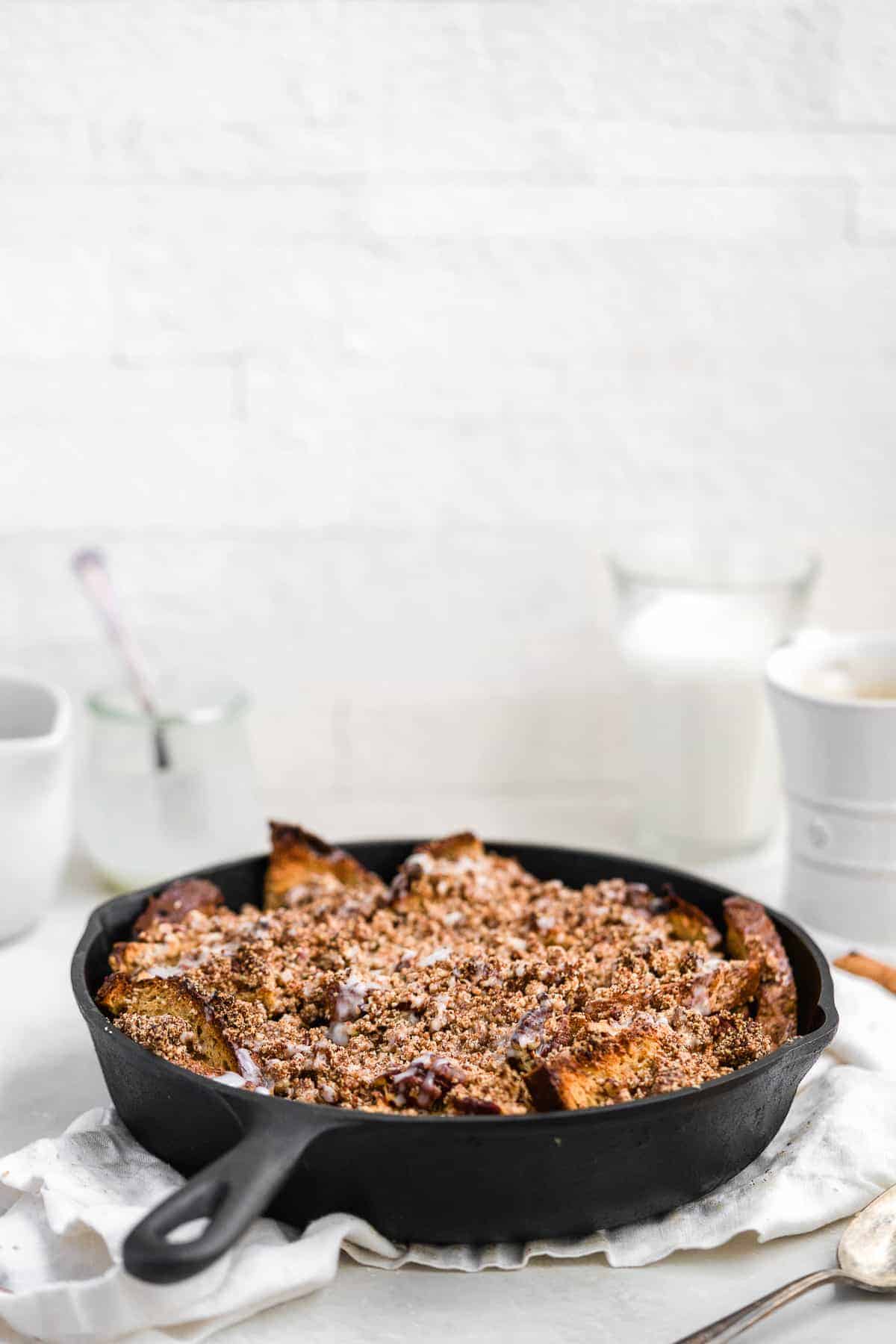 Side view closeup photo of a freshly baked Maple Cinnamon Raisin Dairy-free French Toast Skillet Casserole in a cast iron skillet sitting on a white dish towel on a marble slab. A glass of milk and a coffee cup can be seen in the background.