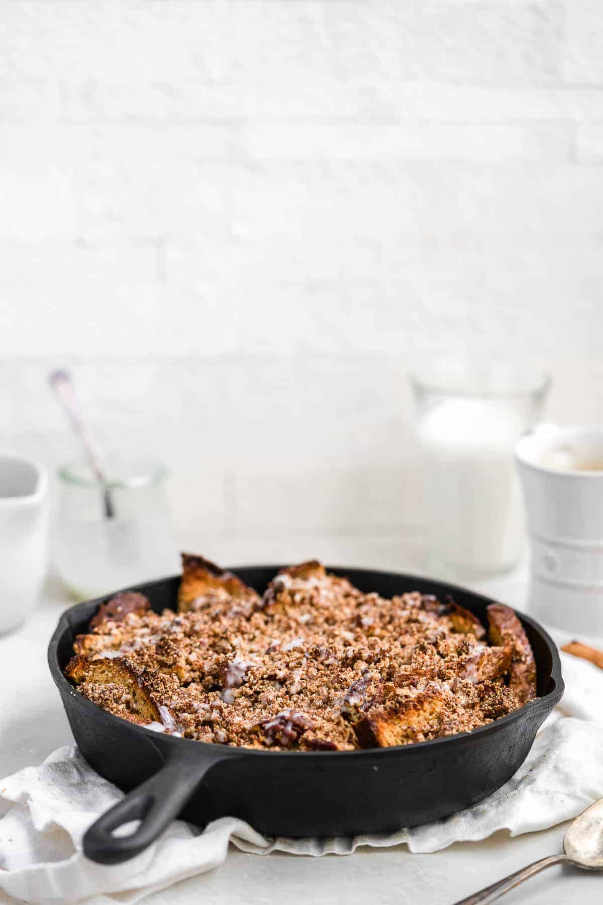 Side view closeup photo of a freshly baked Maple Cinnamon Raisin Dairy-free French Toast Skillet Casserole in a cast iron skillet sitting on a white dish towel on a marble slab.  A glass of milk and a coffee cup can be seen in the background.  