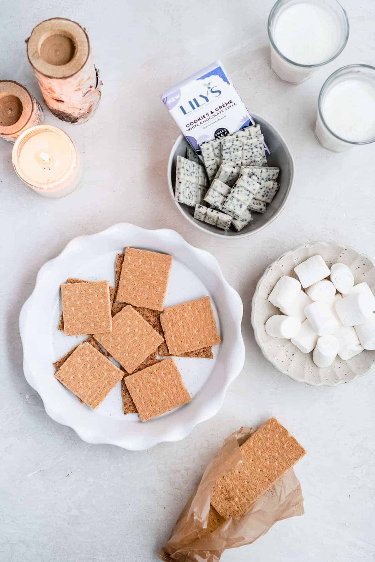 Overhead view of the ingredients that make up this recipe. Graham crackers are on a white plate with scalloped edges. Marshmallows are in a white ceramic bowl and the Lily's chocolate bars are in a white bowl. Two glasses of milk and a candle are in the background.