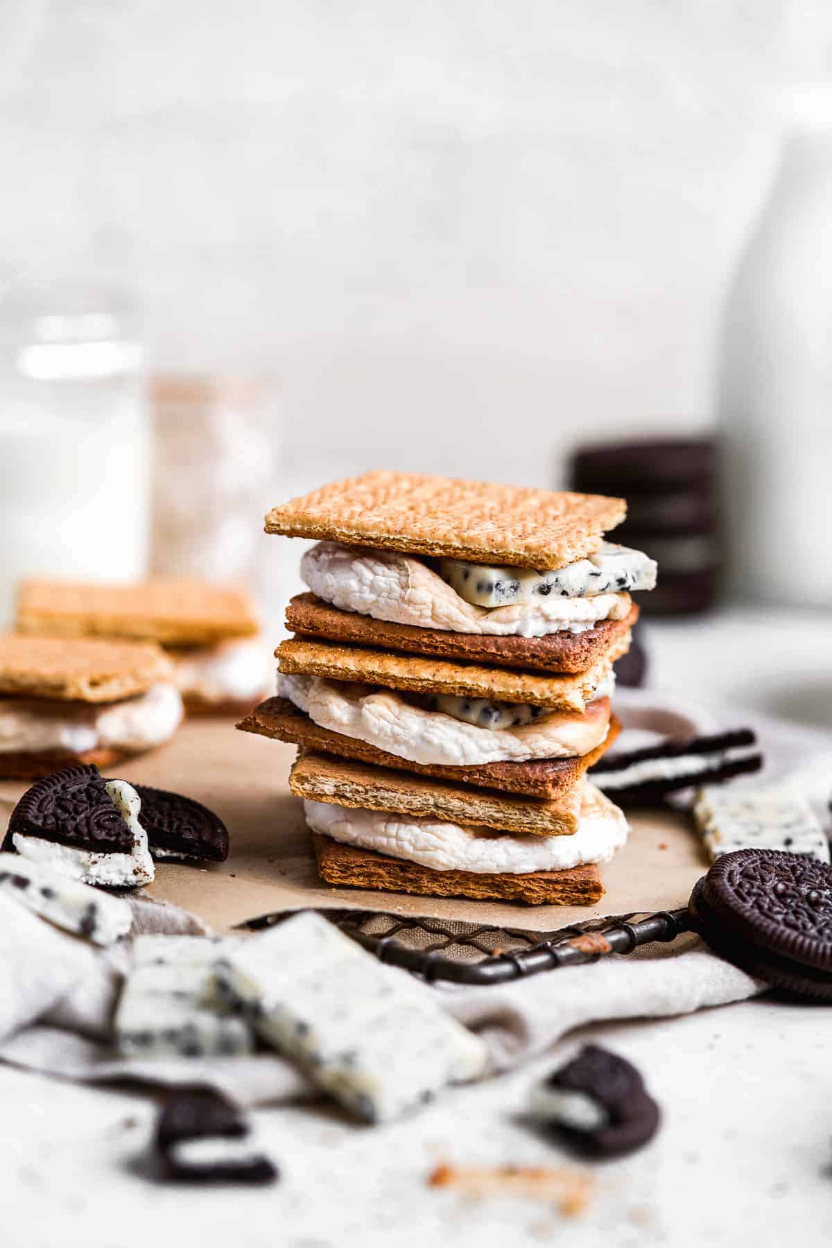 Side view closeup photo of a stack of three Cookies and Cream Gluten-free S'mores sitting on parchment paper on a metal cooling rack. Pieces of Oreo cookies and the Lily's Chocolate bars are sprinkled around.