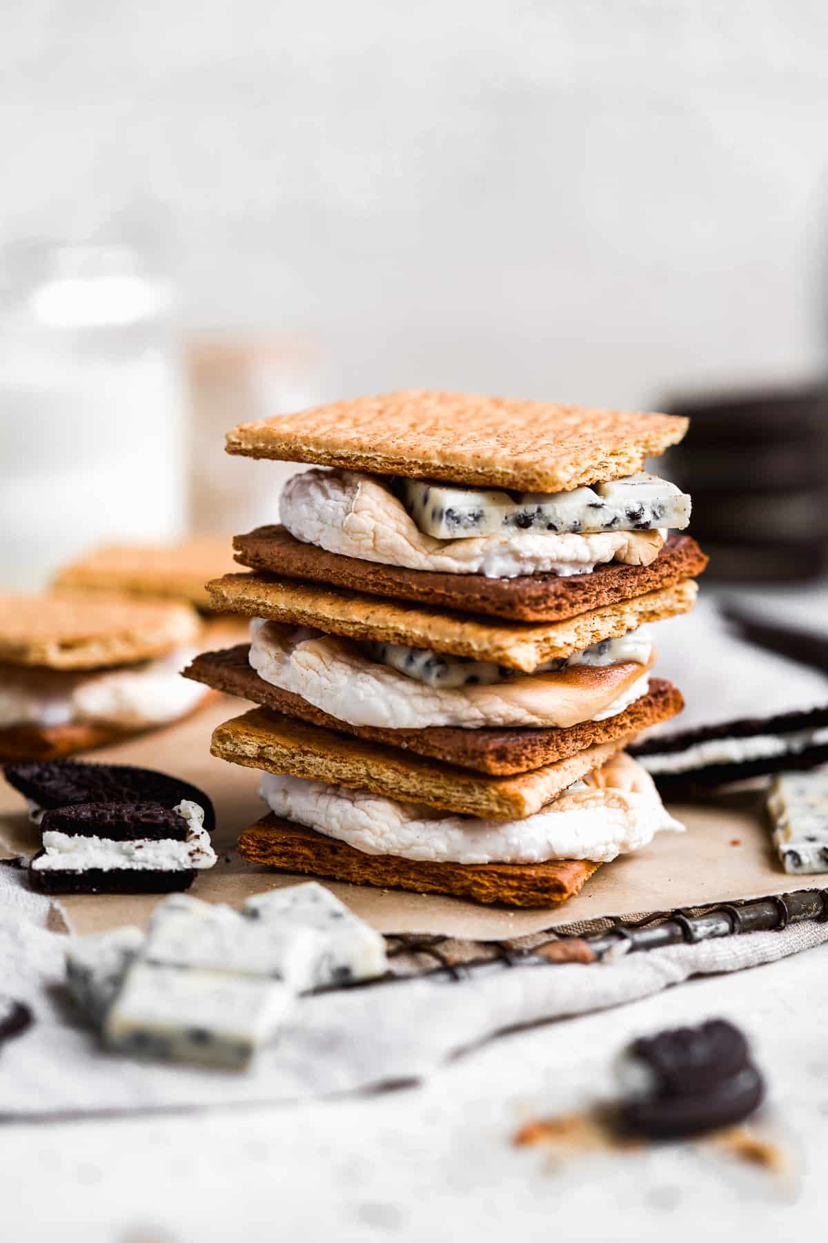 Side view closeup of a stack of 3 Cookies and Cream Gluten-free S'mores  sitting on parchment paper on top of a metal cooling rack.  Additional cookies are in the background and chunks of the chocolate Oreo layer are sprinkled around.  