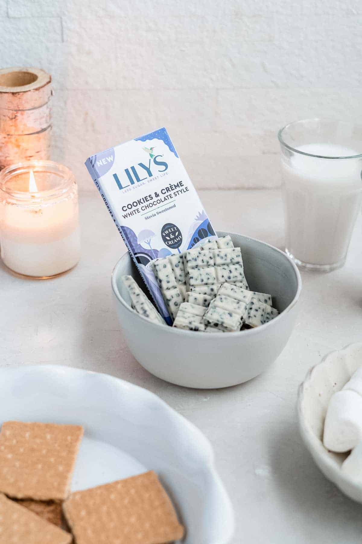A wrapped bar of Lily's White Chocolate Cookies & Creme is sitting in a white ceramic bowl with unwrapped bars. A glass of milk, a candle, a bowl of marshmallows and a plate with graham crackers are in the background.